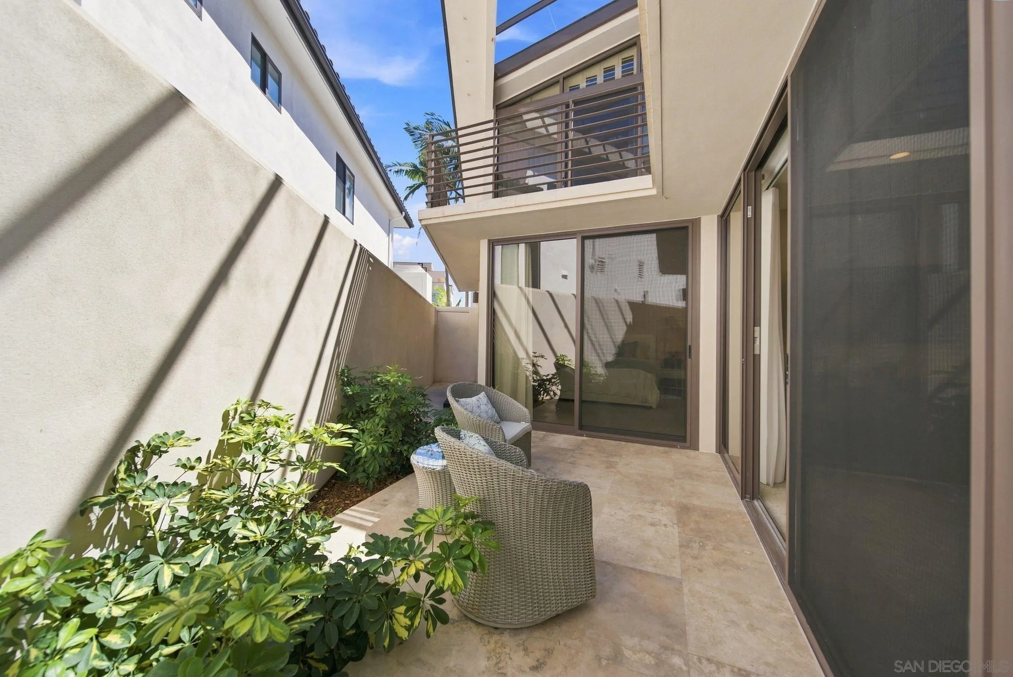 Tiled courtyard patio with wicker chairs, lush planting, sliding glass doors, and an upper balcony.