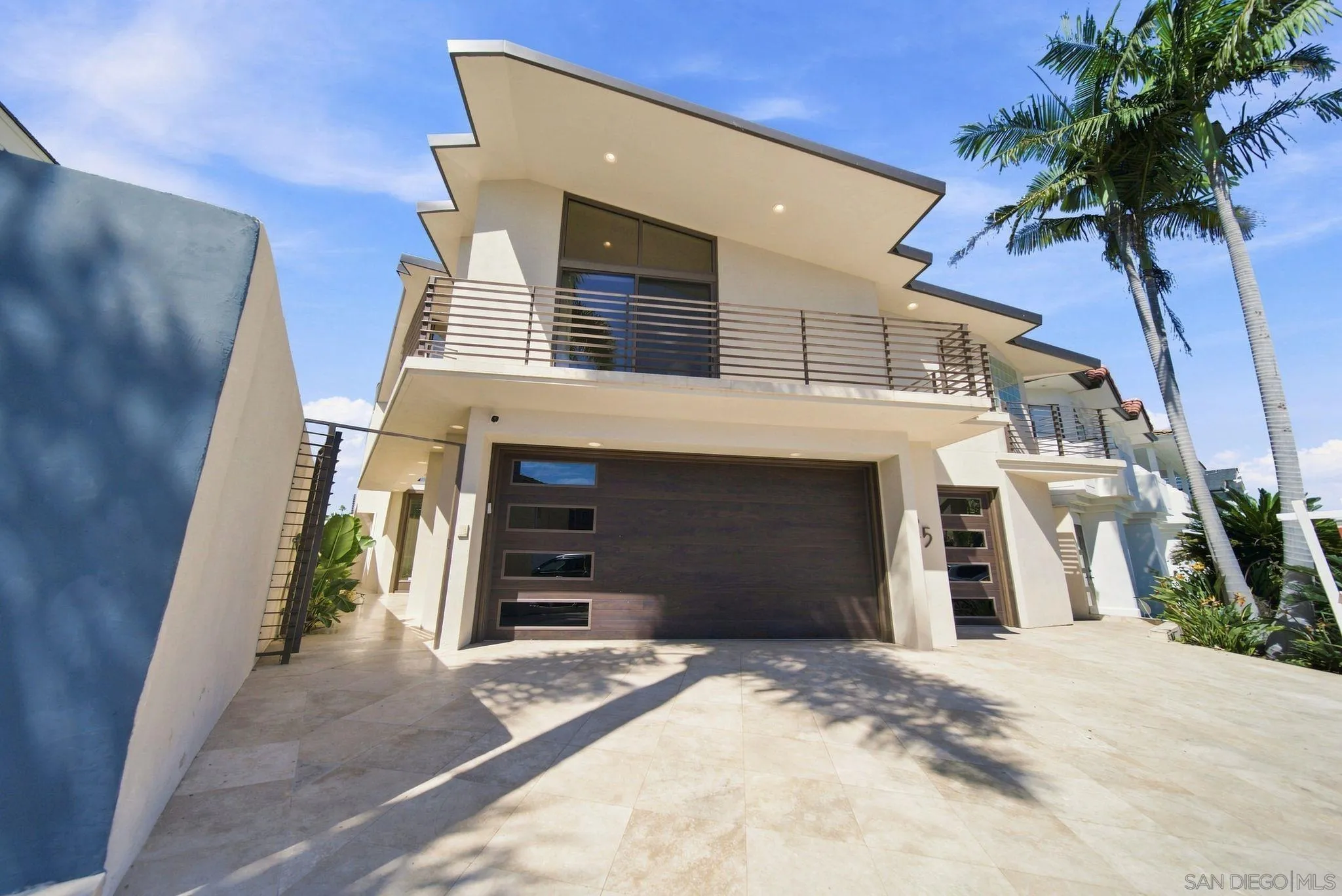 Modern white home with angular rooflines, a wide balcony, dark garage door, and palm trees.