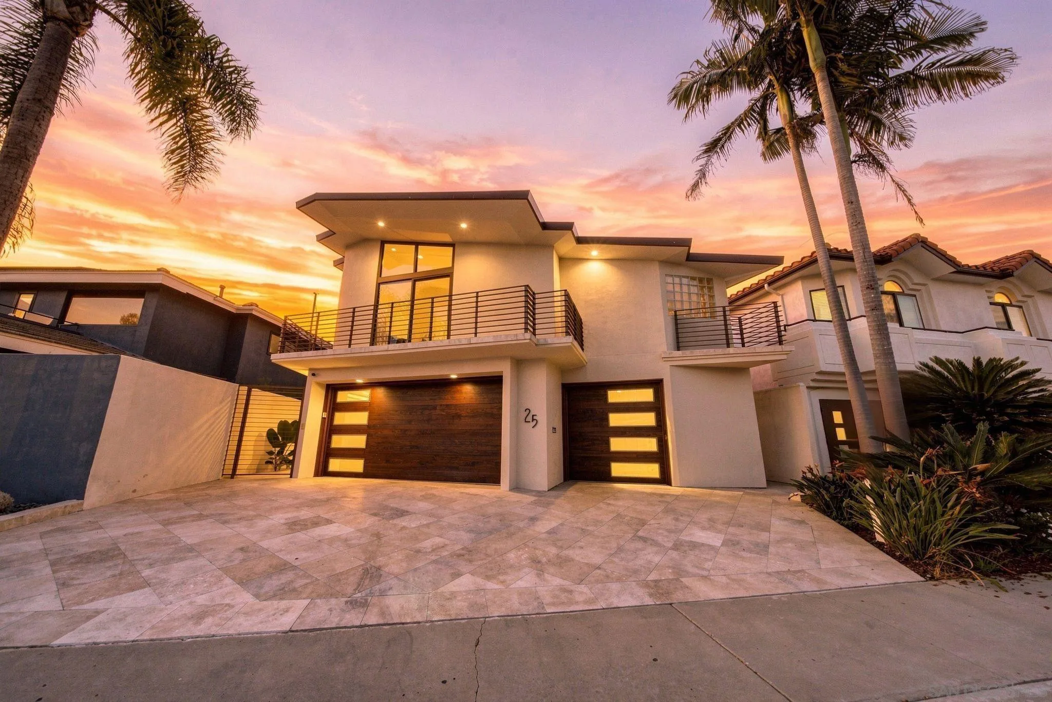 Modern two-story home with balconies and illuminated wood garage doors at sunset, framed by palm trees.