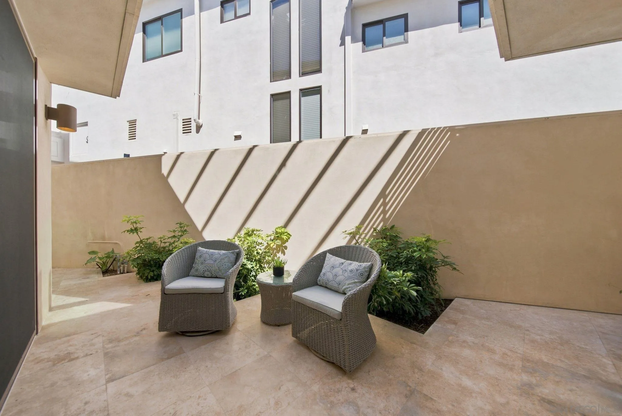 Private courtyard patio with wicker chairs, planted shrubs, and sharp diagonal shadows.
