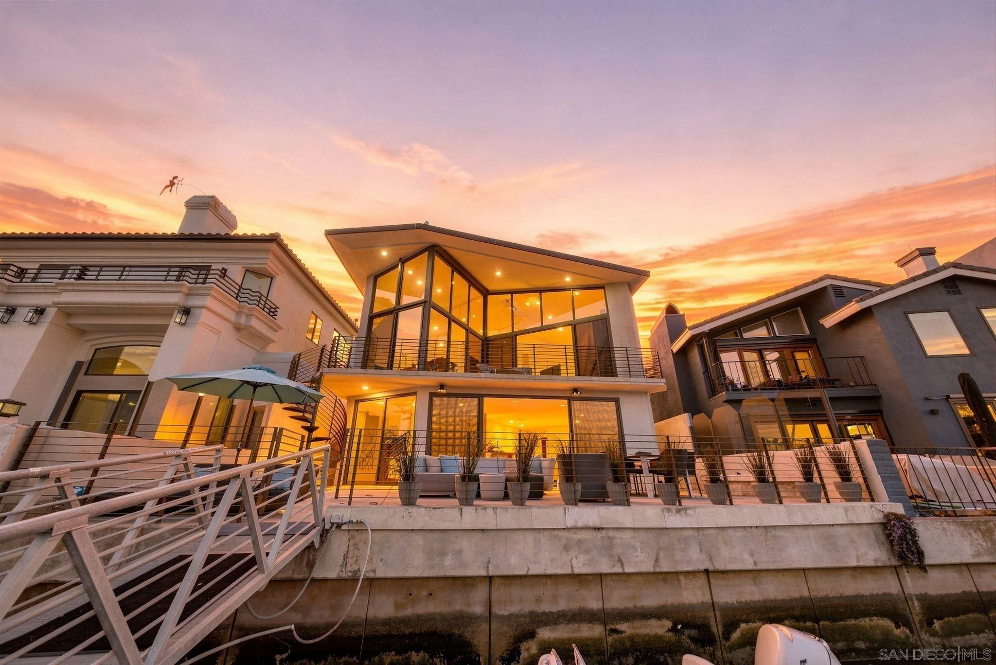 Modern waterfront home with a glass facade and terrace glowing at sunset.