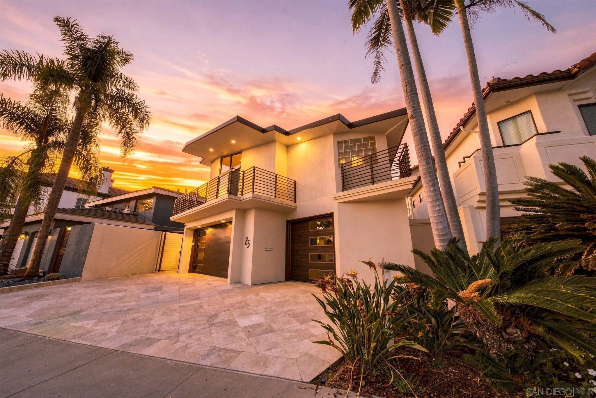 Modern two-story home with palm trees, tropical landscaping, and a glowing sunset.