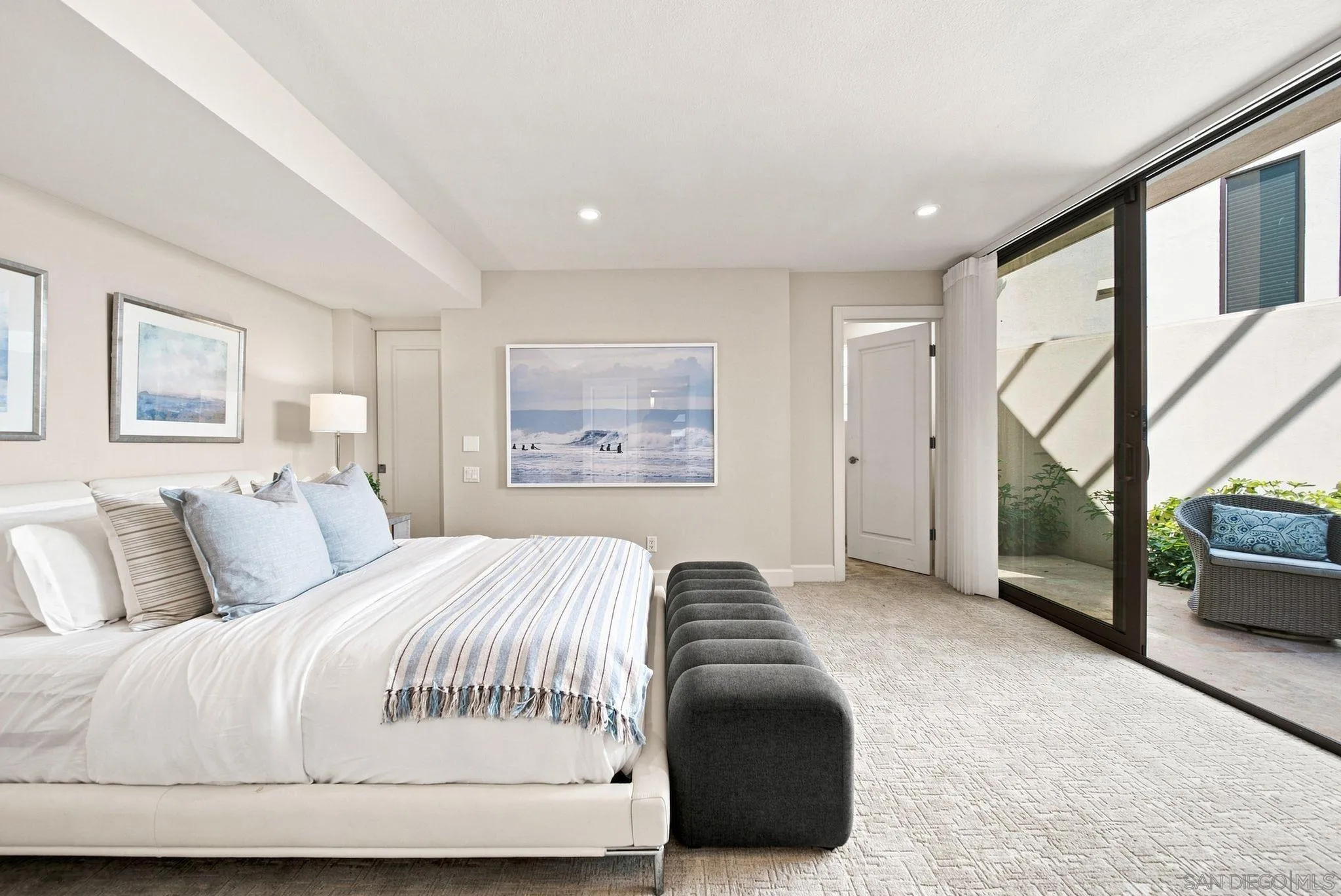 Bright bedroom with a white bed, gray bench, coastal artwork, and sliding glass doors to a planted patio.