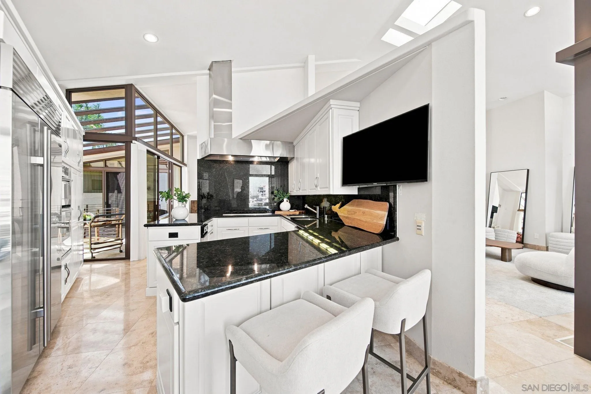 Modern white kitchen with black granite counters, breakfast bar seating, and large windows opening to a patio.