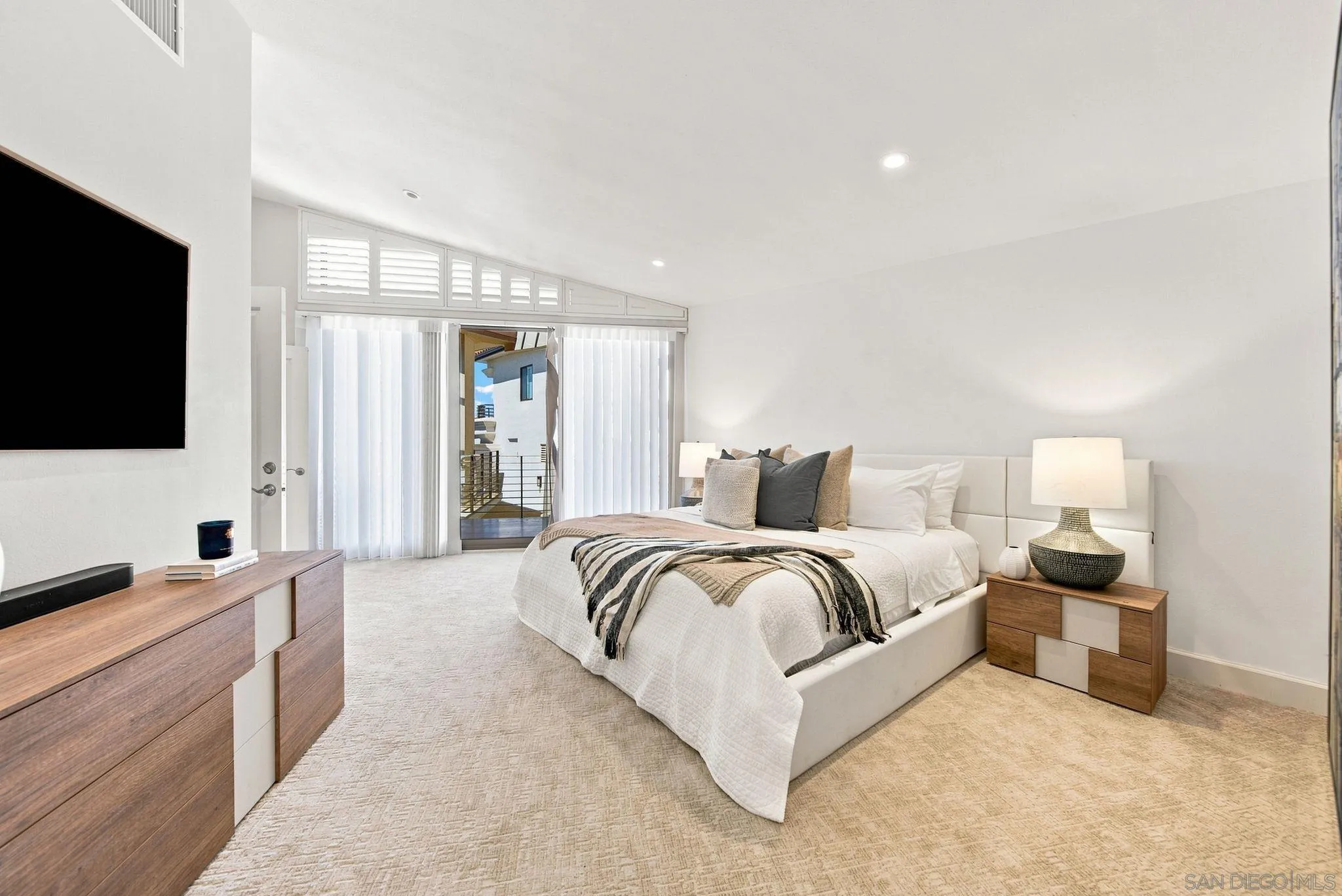 Furnished bedroom with a white bed, wood furniture, and sliding glass doors with vertical blinds.