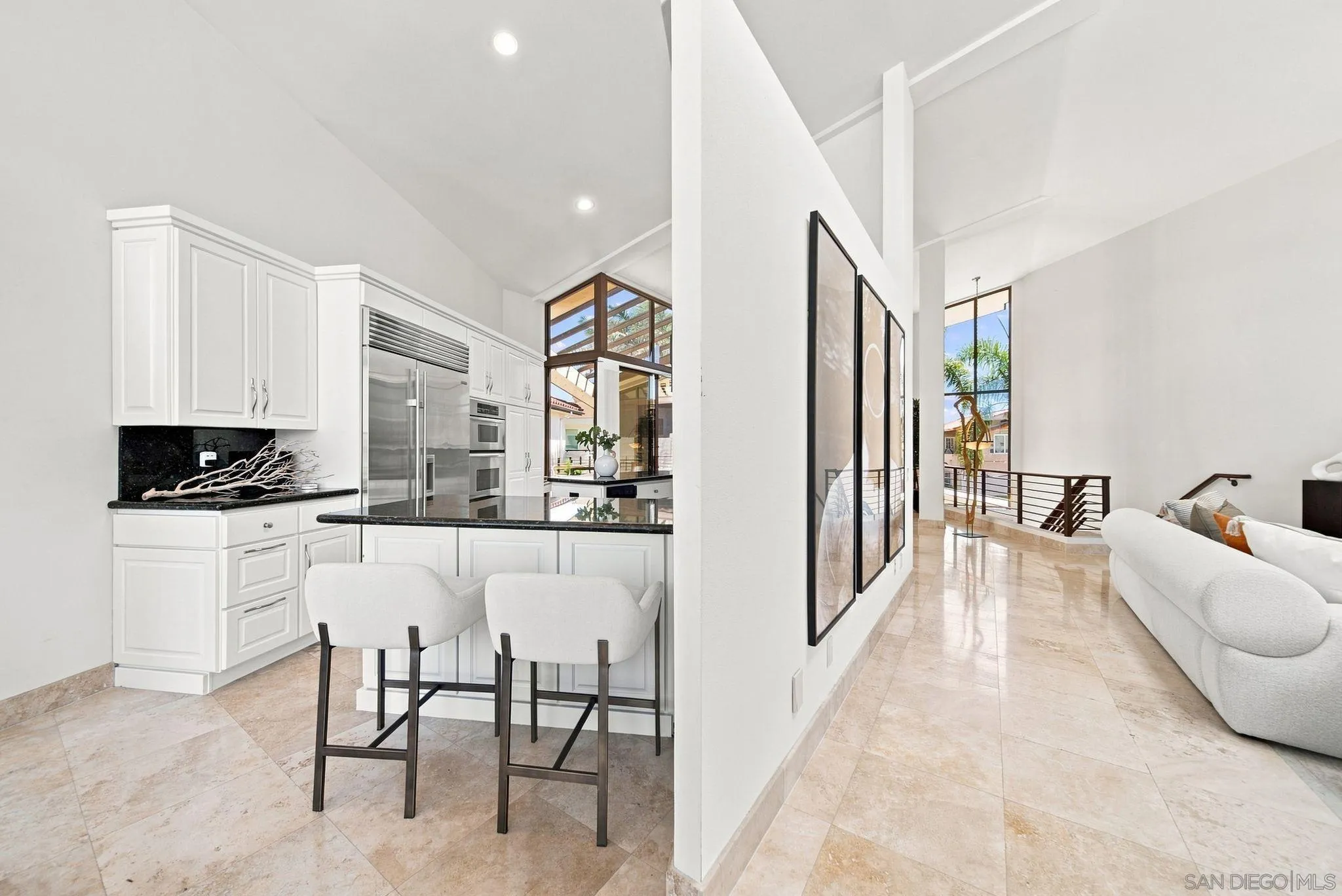 Open kitchen with white cabinetry and black island beside a bright living area with tall windows.