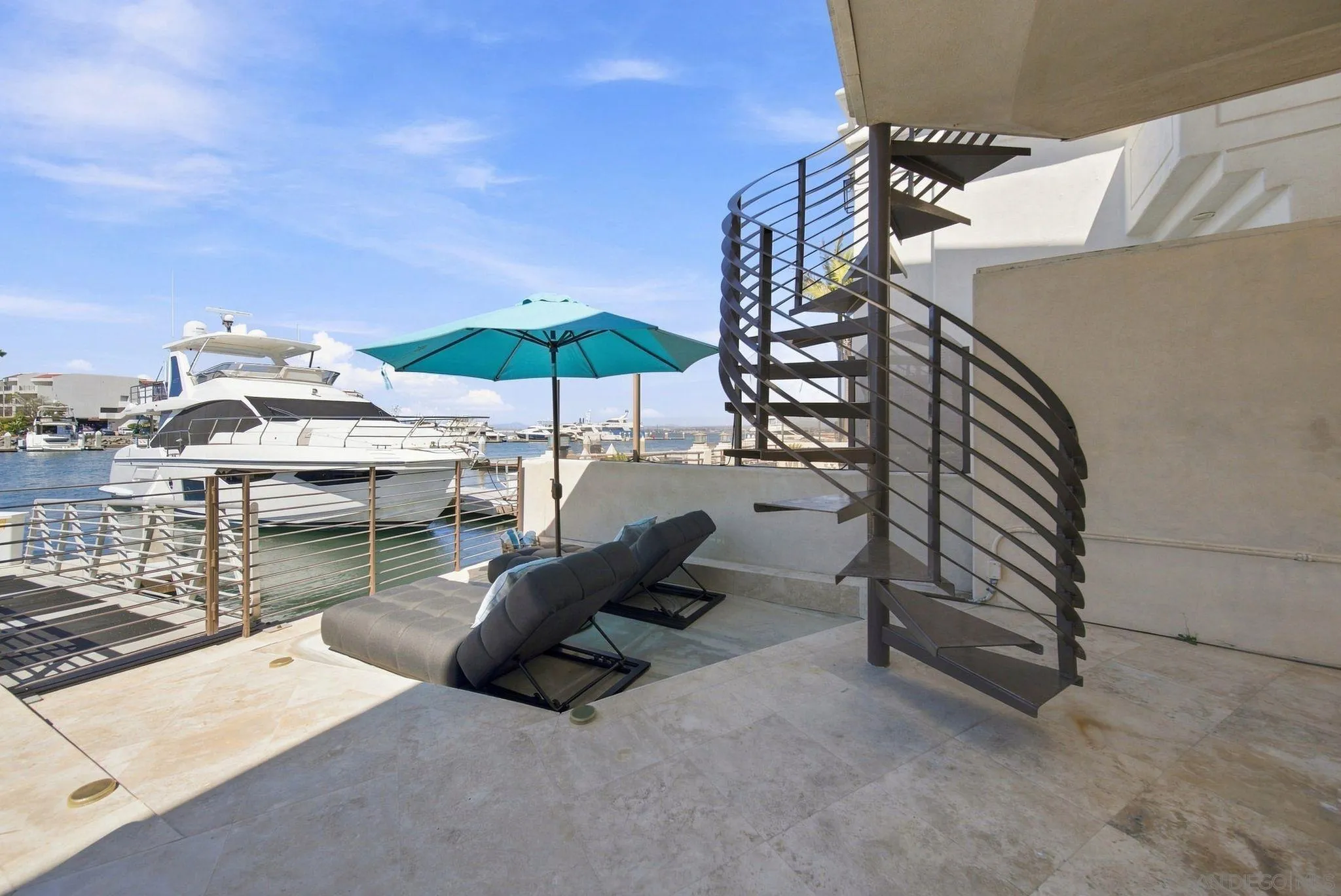Waterfront terrace with reclining loungers, a turquoise umbrella, and a curved staircase beside a marina.