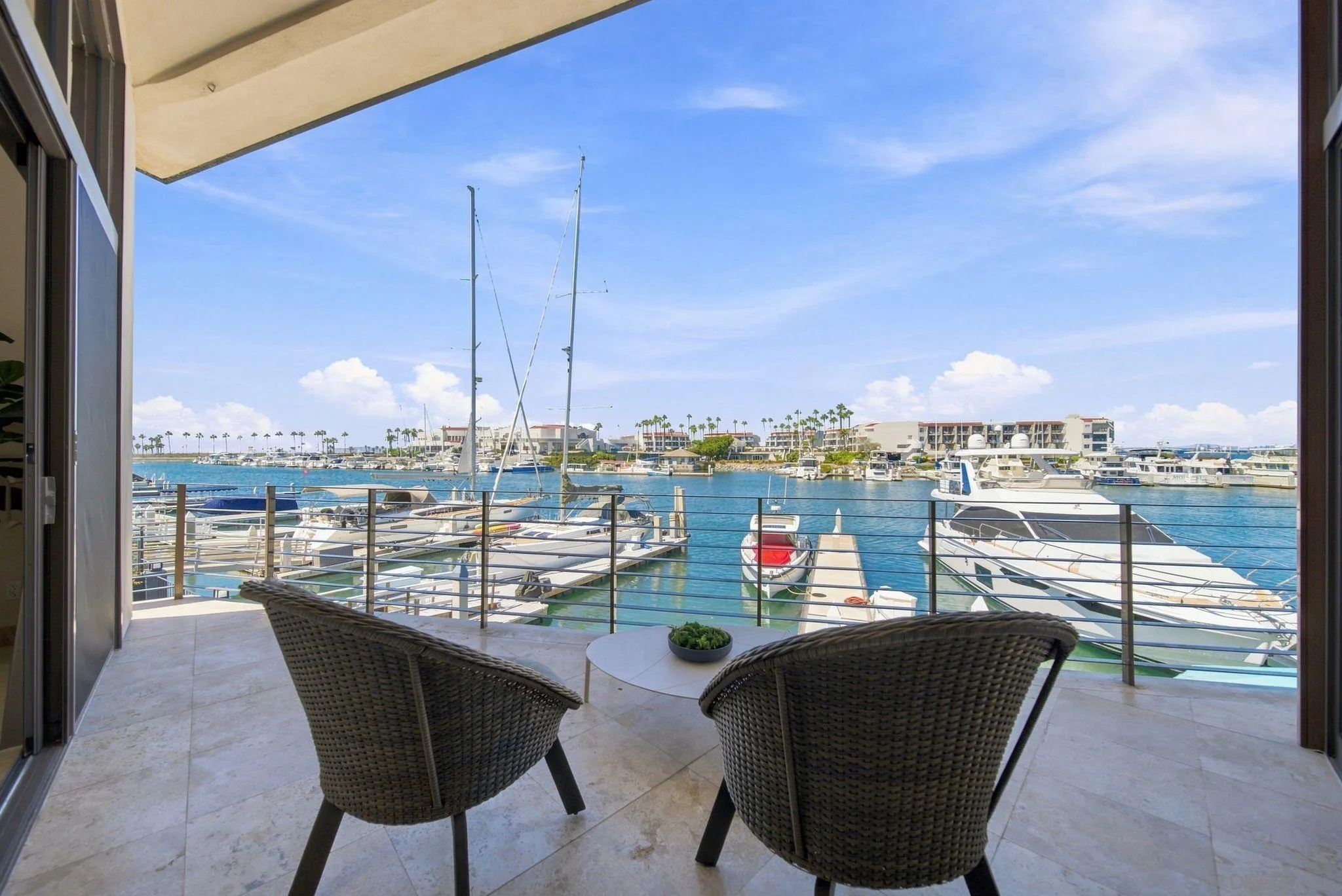 Furnished balcony overlooking a marina, with two woven chairs and boats moored just beyond the railing.