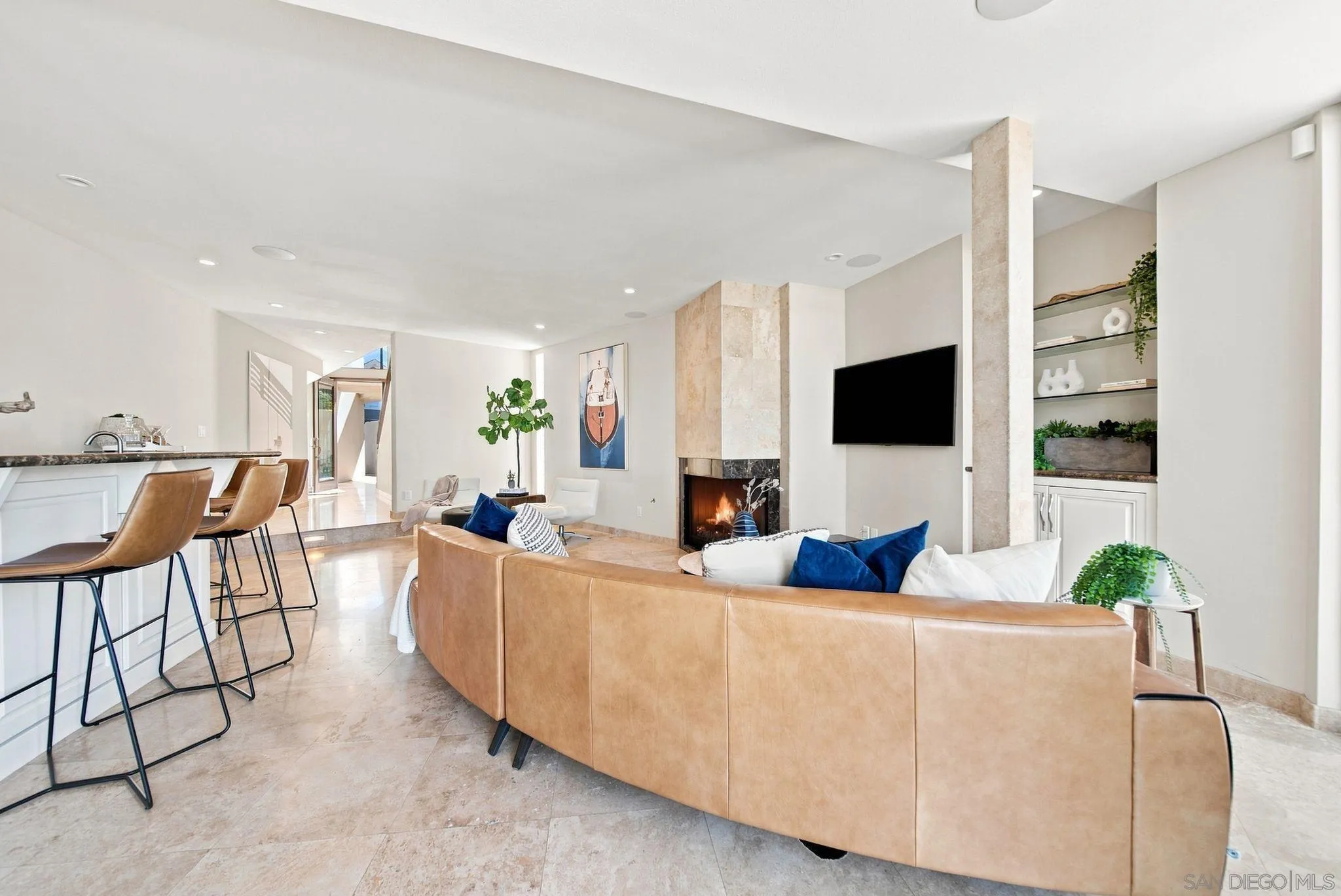 Open-plan living room with a curved tan leather sofa, lit stone fireplace, and kitchen island with bar stools.
