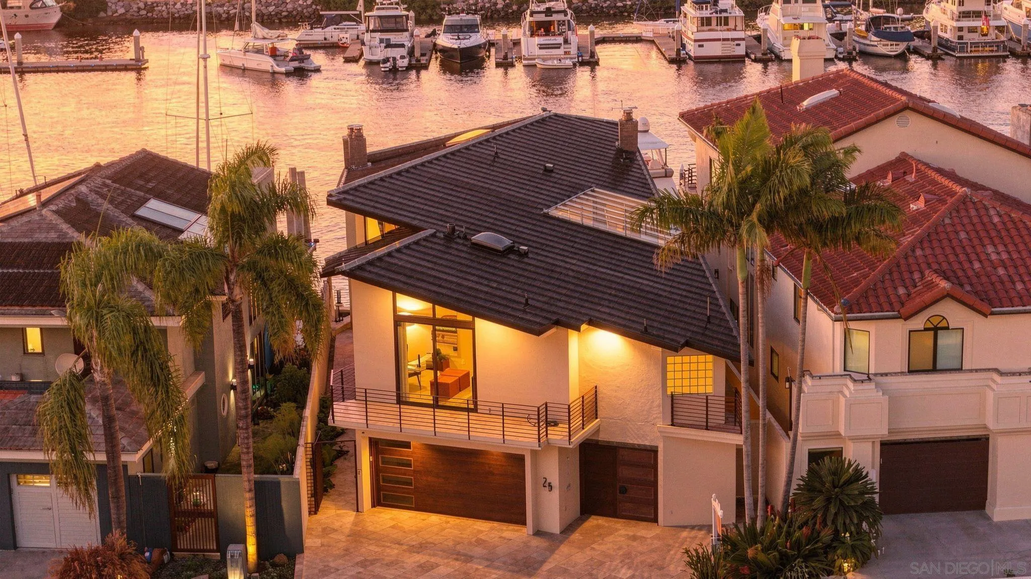 Waterfront home with palm trees and balconies overlooking a marina filled with boats at sunset.