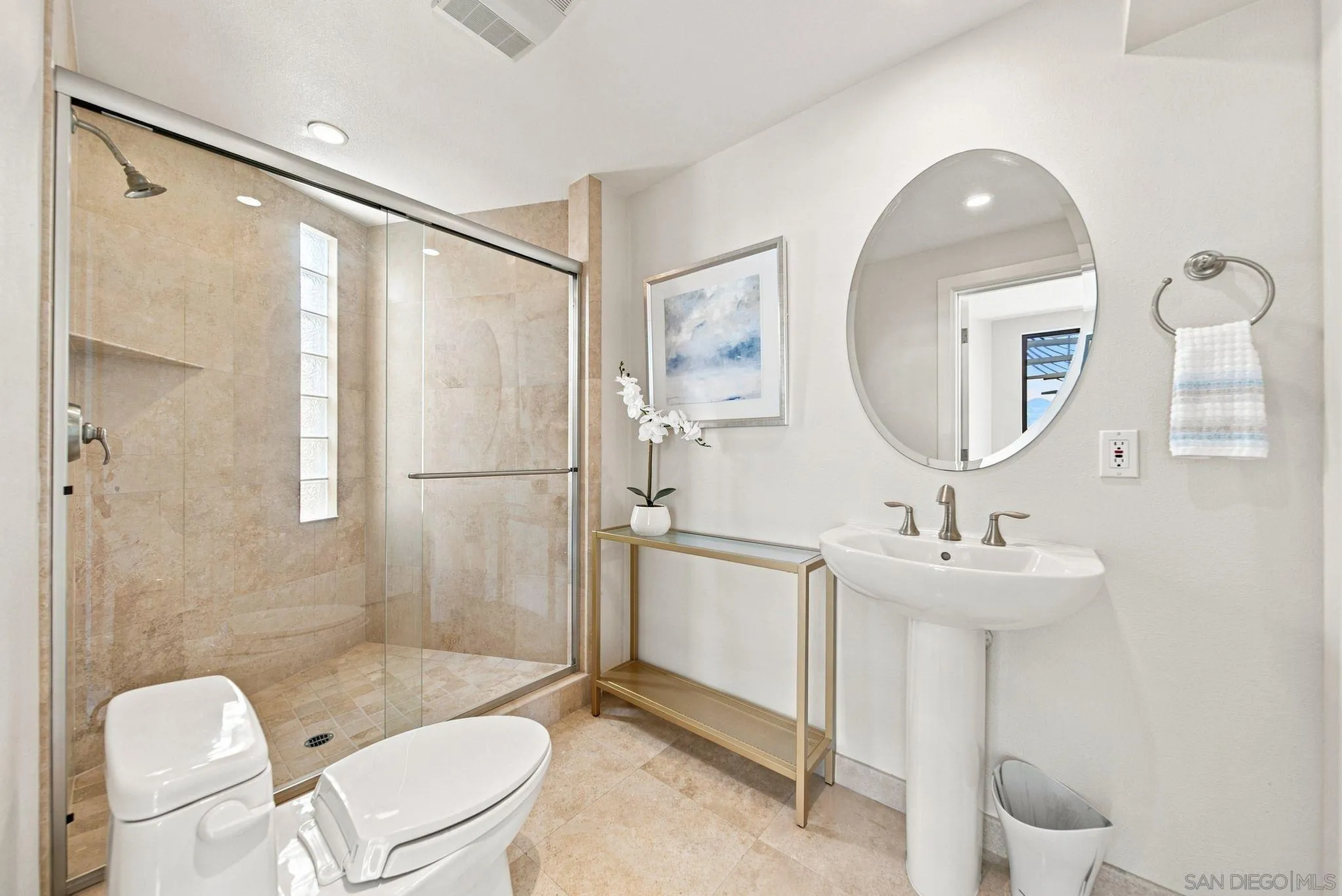 Bathroom with a beige stone-tile glass shower, pedestal sink, and large oval mirror.