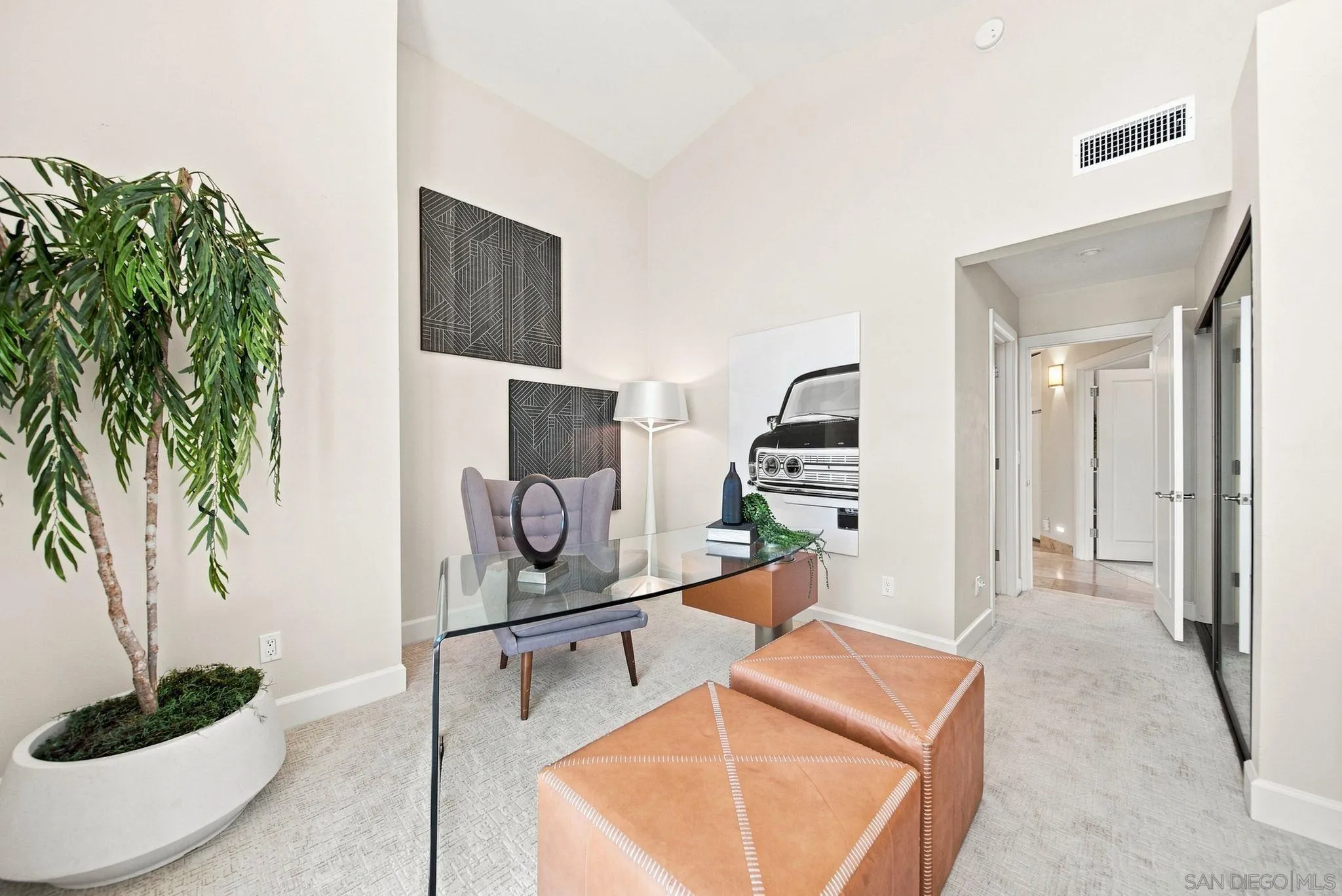 Staged sitting area with a glass desk, lavender chair, tan ottomans, and a large potted tree.