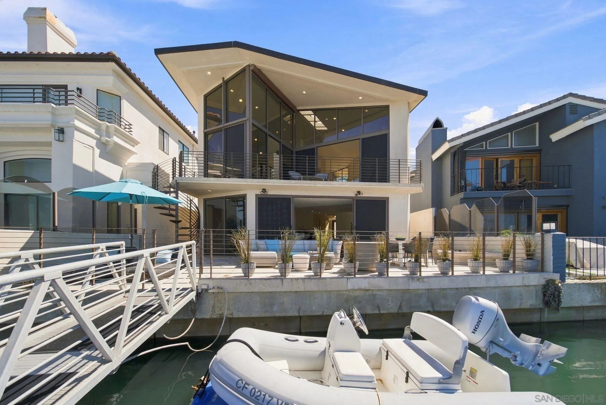 Modern waterfront home with expansive glass walls, furnished terraces, and a dock with an inflatable boat.