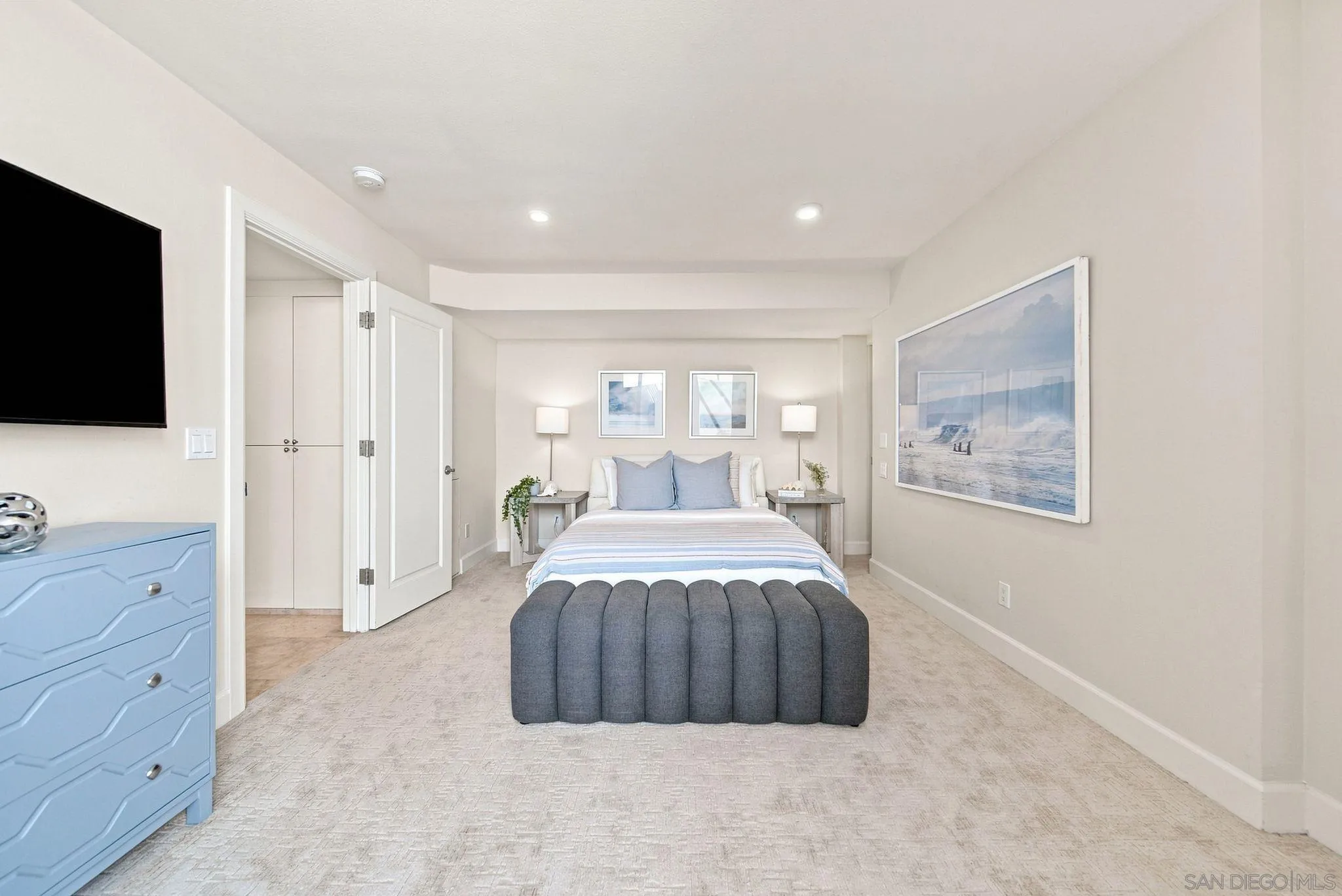 Staged bedroom with a centered bed, gray upholstered bench, and blue dresser.