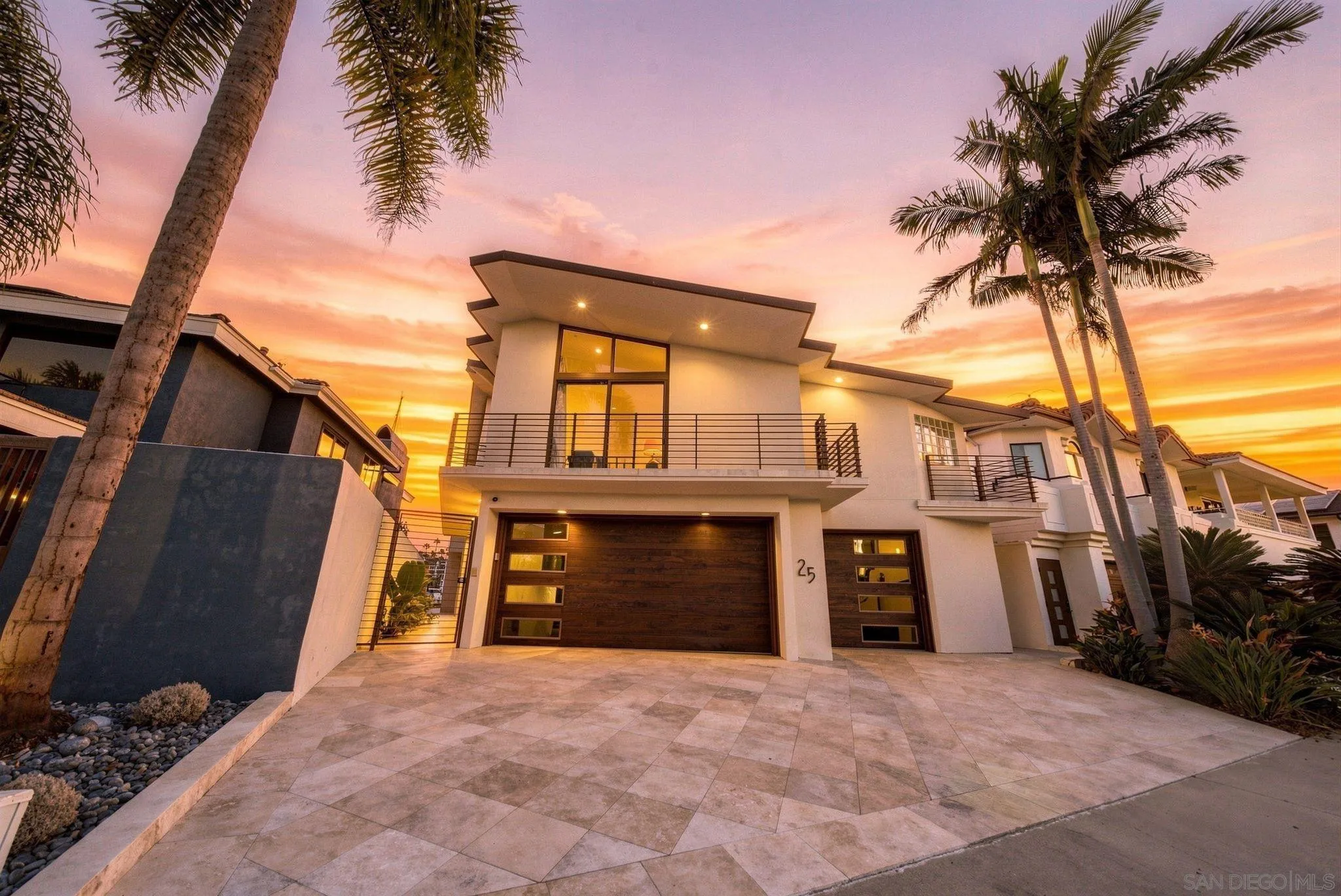 Modern white two-story home with a tiled driveway, wood garage door, and palm trees at sunset.