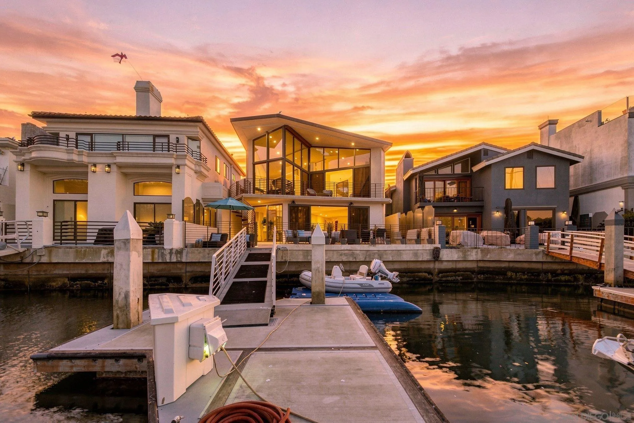 Modern glass-fronted waterfront home at sunset, viewed from its dock with boats moored alongside.
