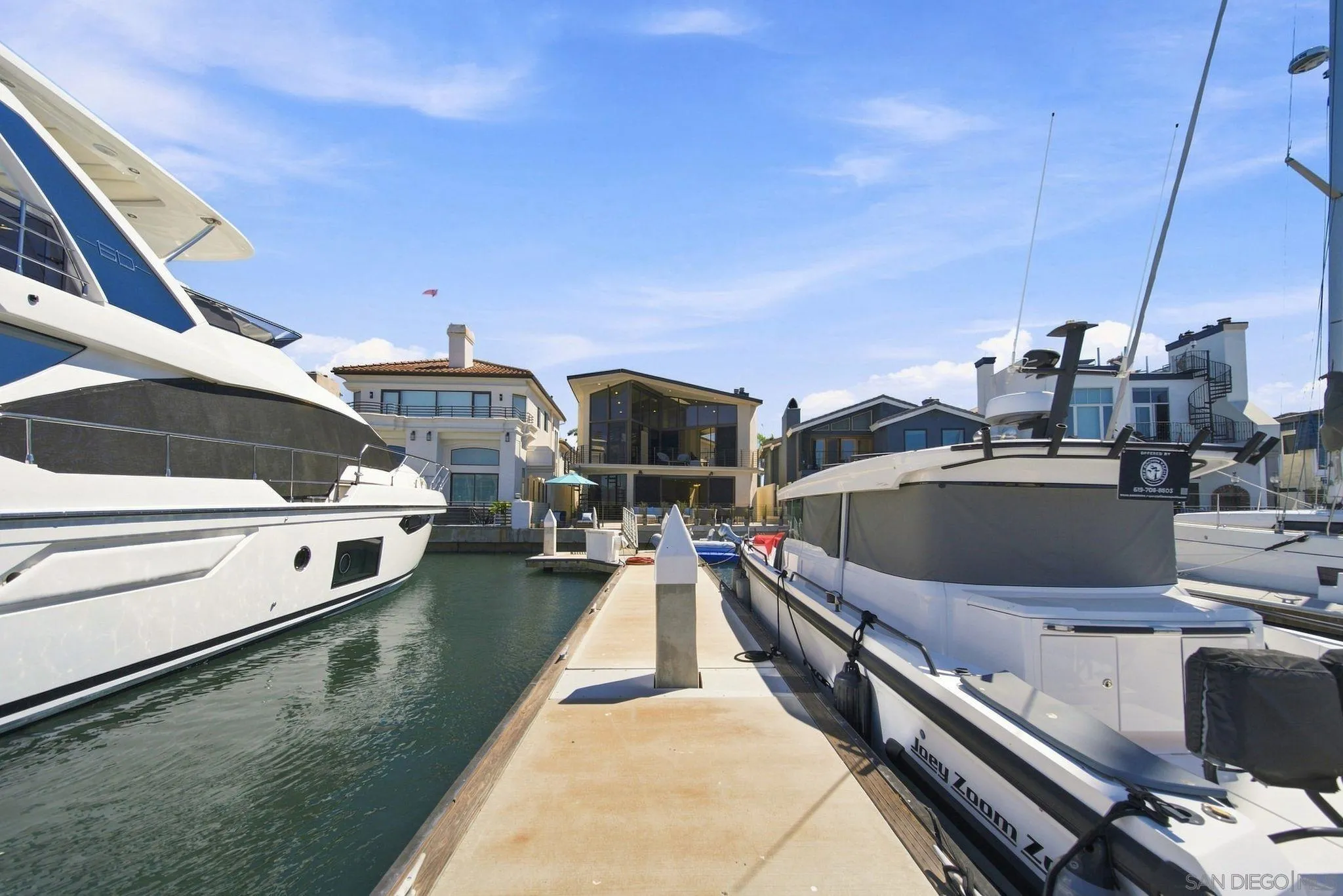 Marina dock between yachts, with modern waterfront homes in the background.