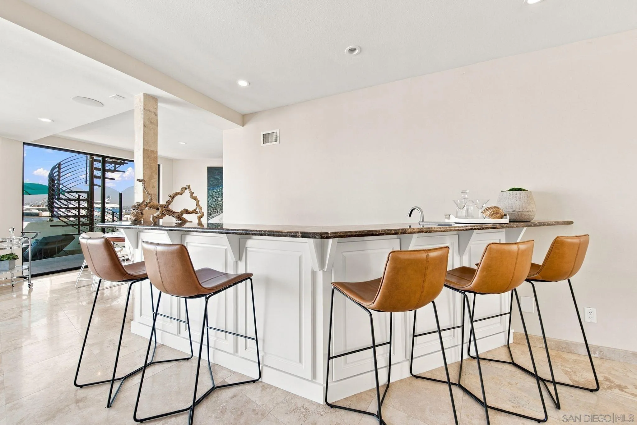 Kitchen breakfast bar with a dark stone countertop, white island, and tan leather stools beside glass doors.