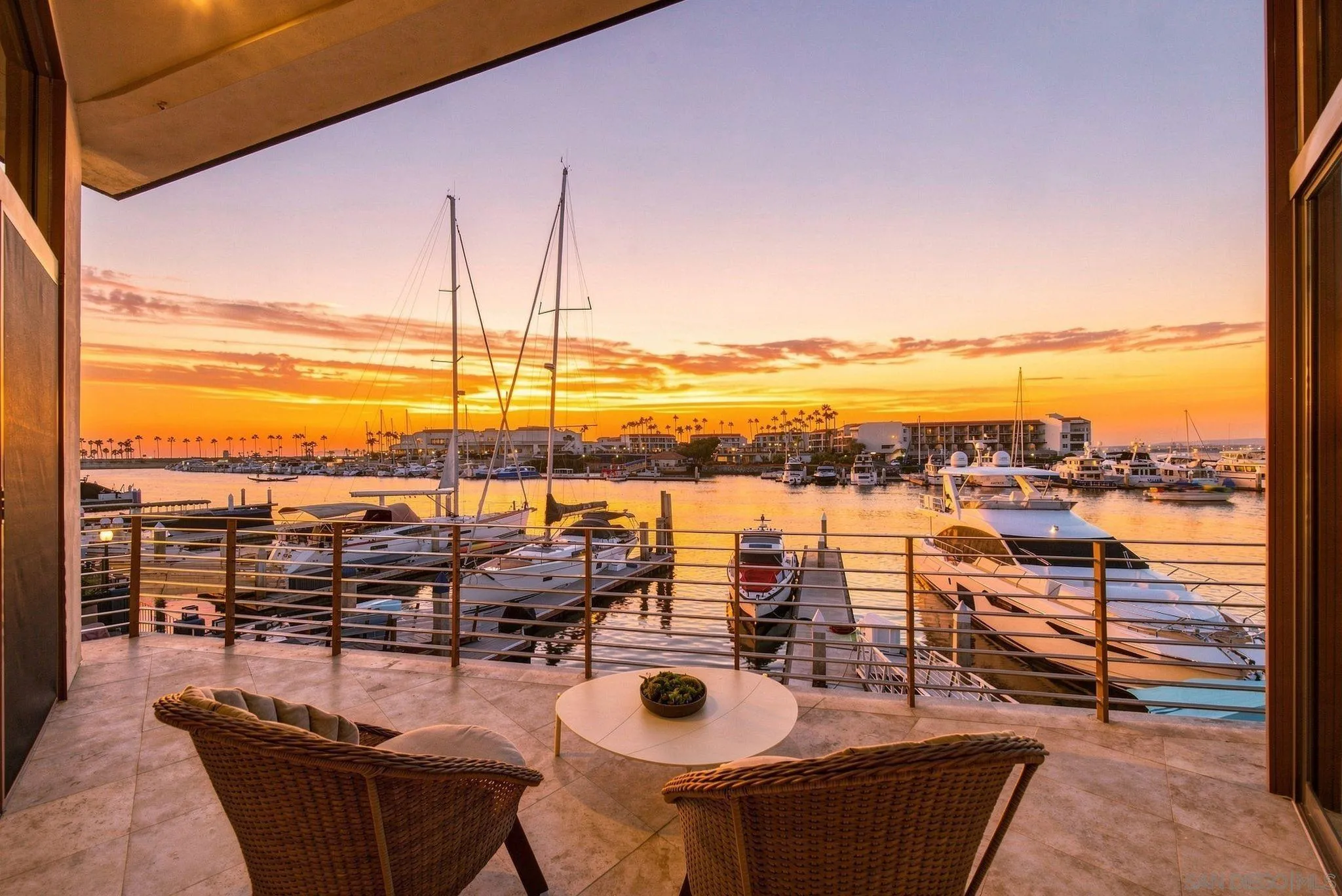 Furnished terrace overlooking a marina at sunset, with wicker chairs and boats moored below.