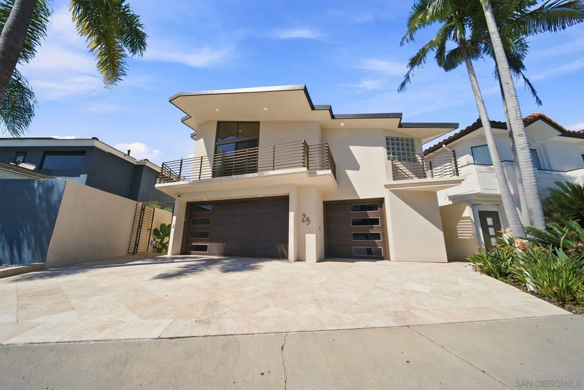 Modern two-story home with angular rooflines, balcony railings, dark garage doors, and palm trees.