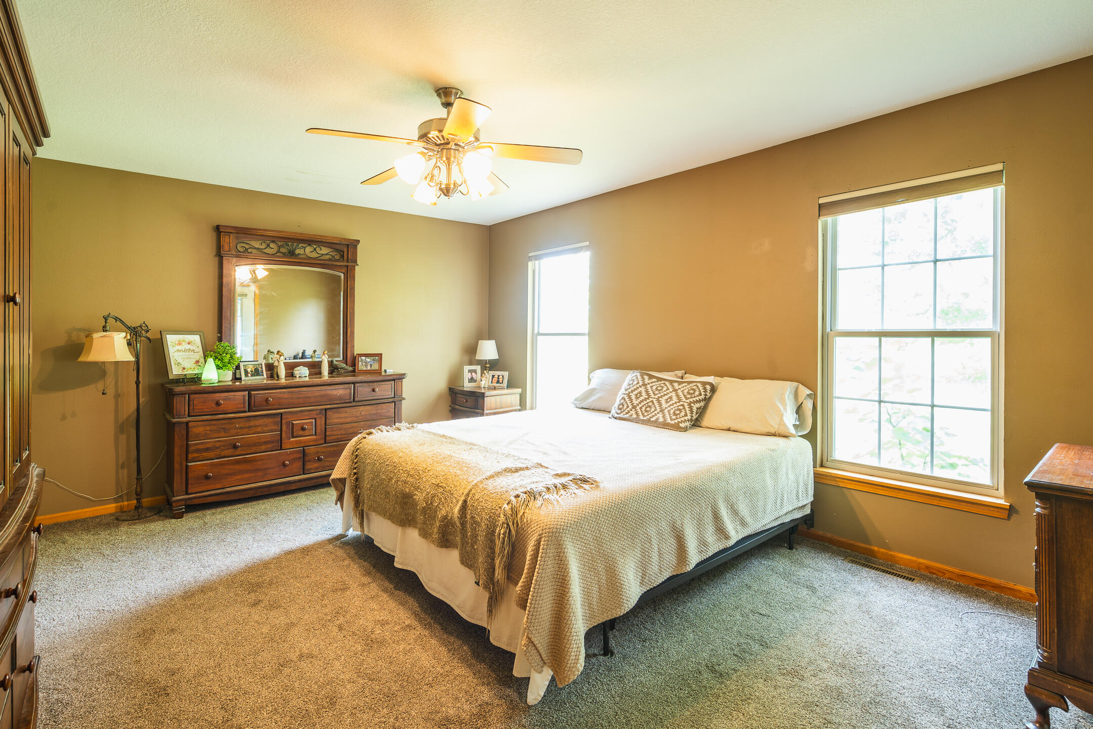 Furnished bedroom with a large bed, dark wood dresser, decorative mirror, and two windows.