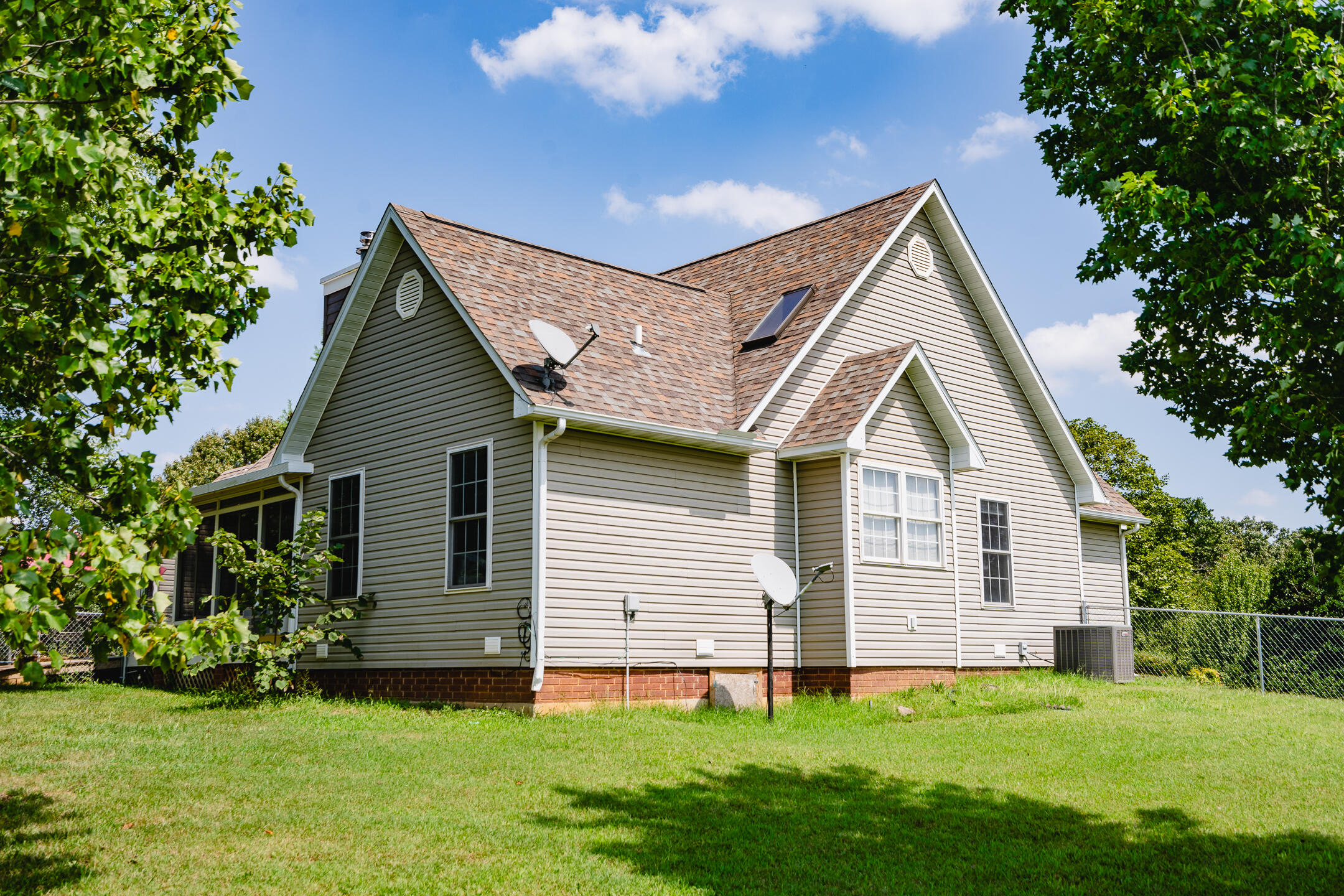 Side view of a light-colored house with a multi-gabled shingle roof, satellite dishes, and a broad green lawn.