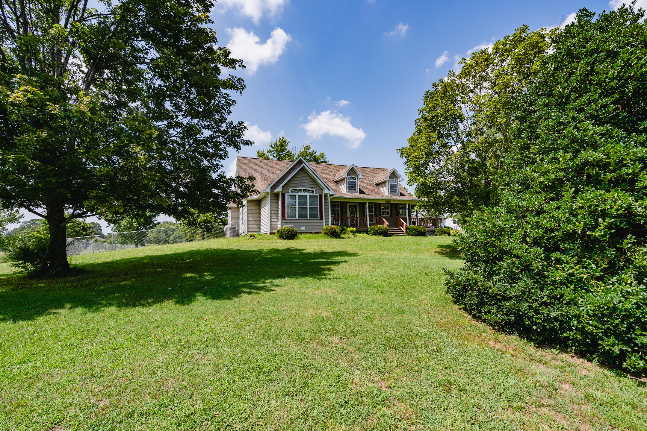 Two-story home with a covered porch and dormers beyond a broad, tree-framed lawn.