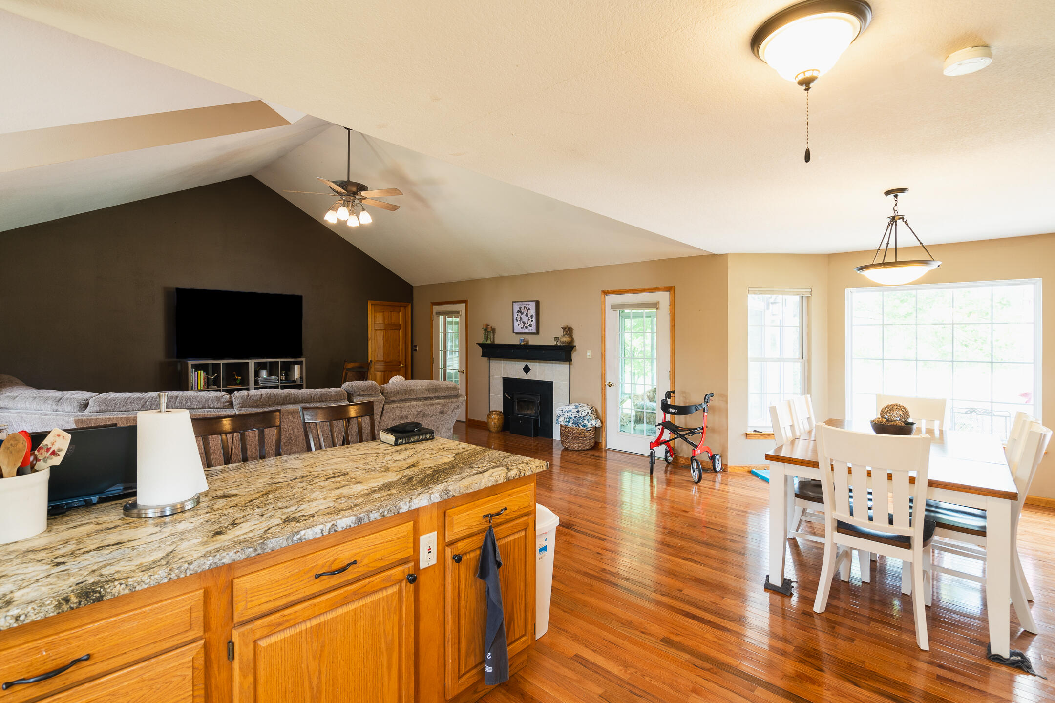 Open-plan living and dining room with vaulted ceiling, polished hardwood floors, granite island, and fireplace.