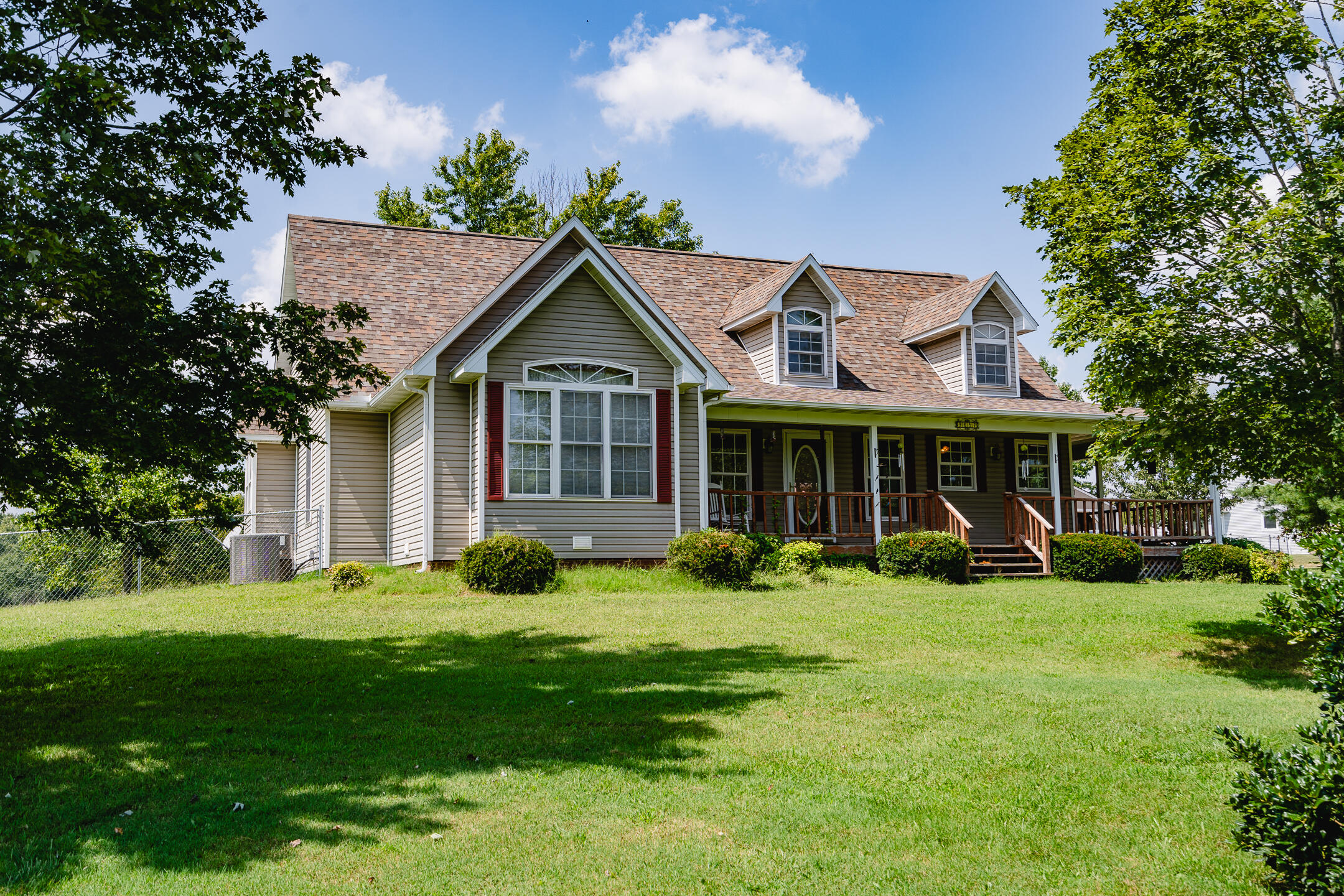 Two-story beige house with dormer windows and a long covered porch, framed by trees above a broad lawn.