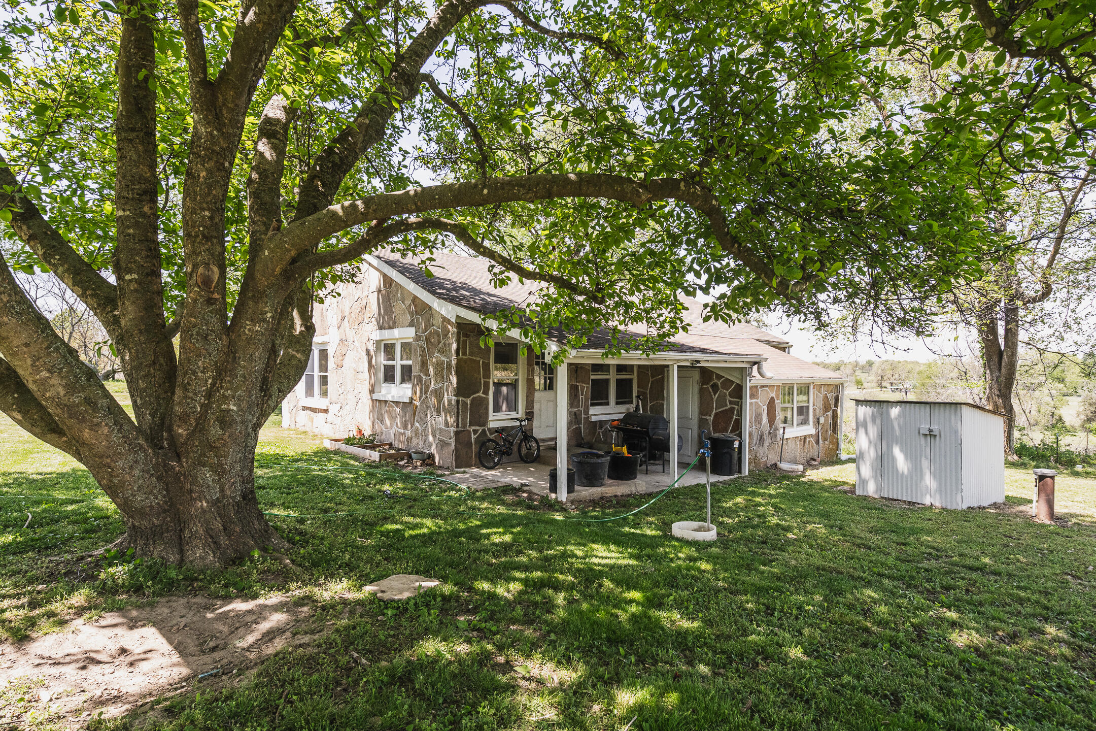 Stone house with a covered patio and shed beneath a large leafy tree.