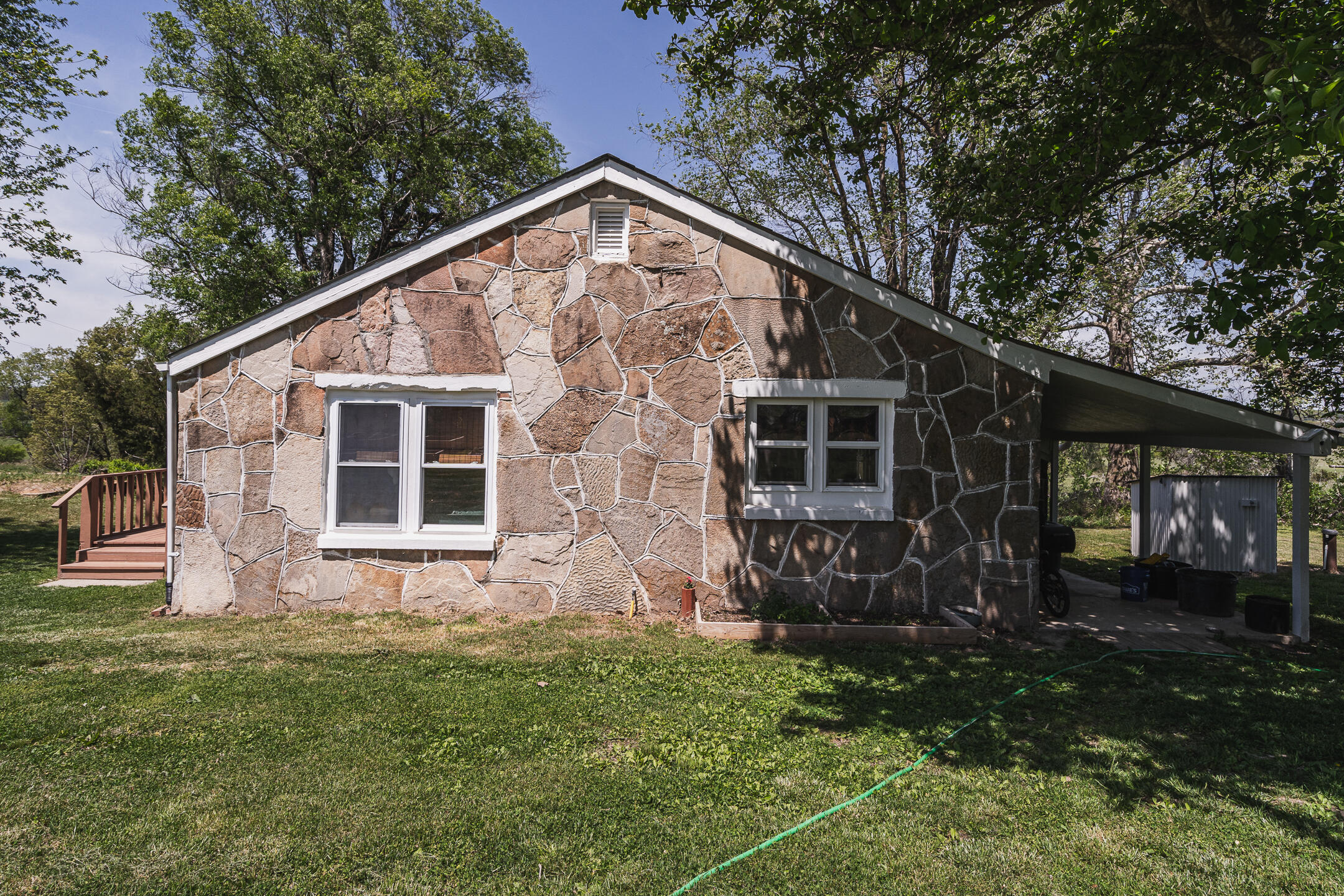 Stone-faced single-story home with a side carport and wooden deck on a tree-shaded lawn.