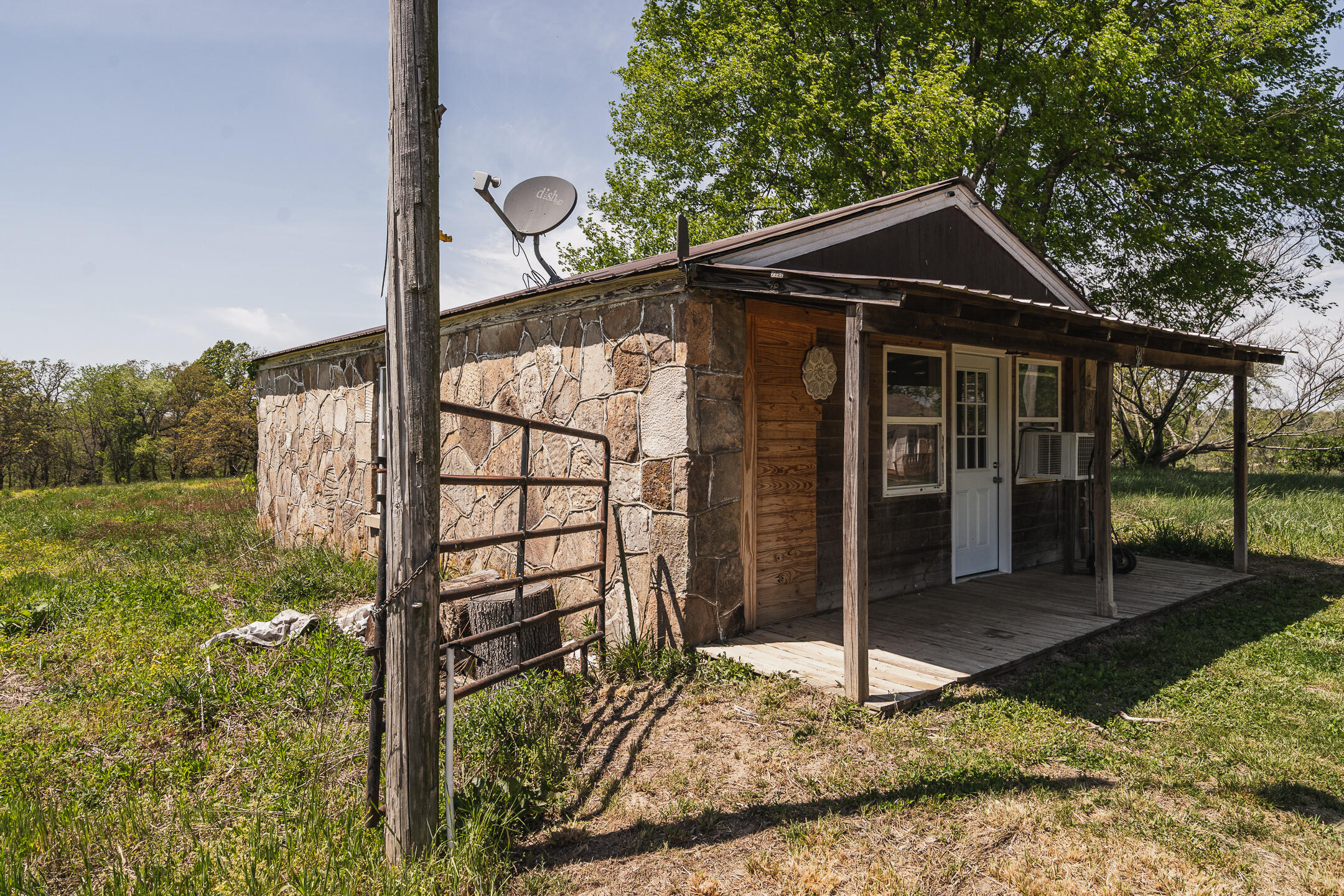 Rustic stone-and-wood cabin with a covered porch, satellite dish, and metal gate in a grassy rural setting.