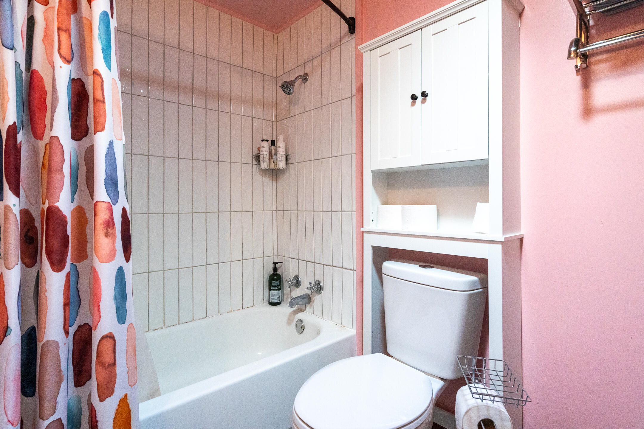 Pink bathroom with a white bathtub, colorful shower curtain, and cabinets above the toilet.