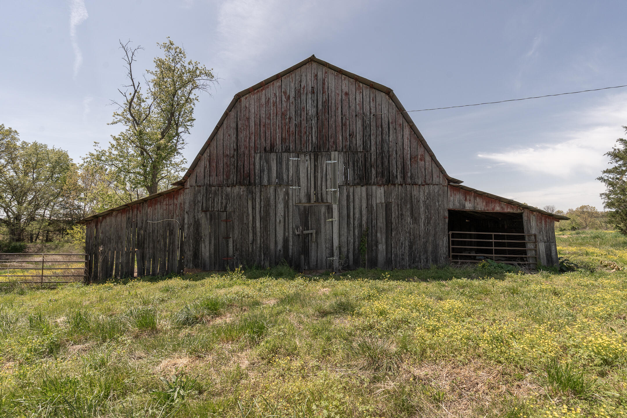 Weathered gambrel-roof barn with an open side bay amid a grassy, flower-dotted field.