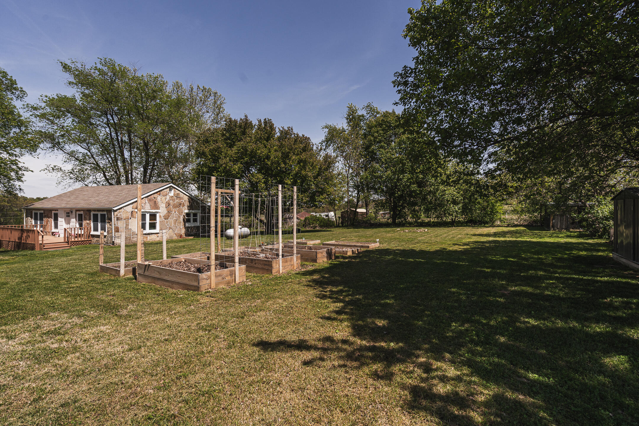 Stone-sided home and wooden deck beside raised garden beds in a large tree-shaded backyard.