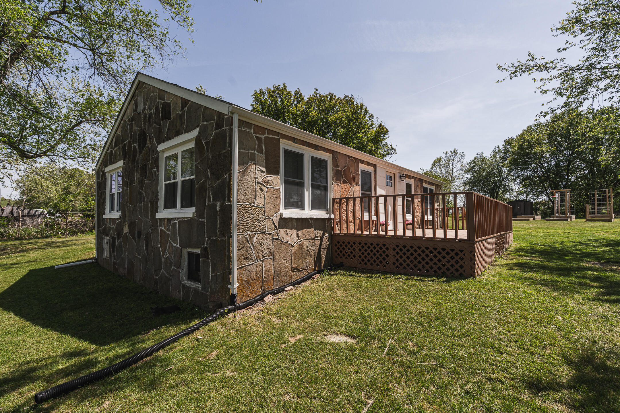 Stone-sided home with a raised wooden deck and garden structures on a sunny lawn.