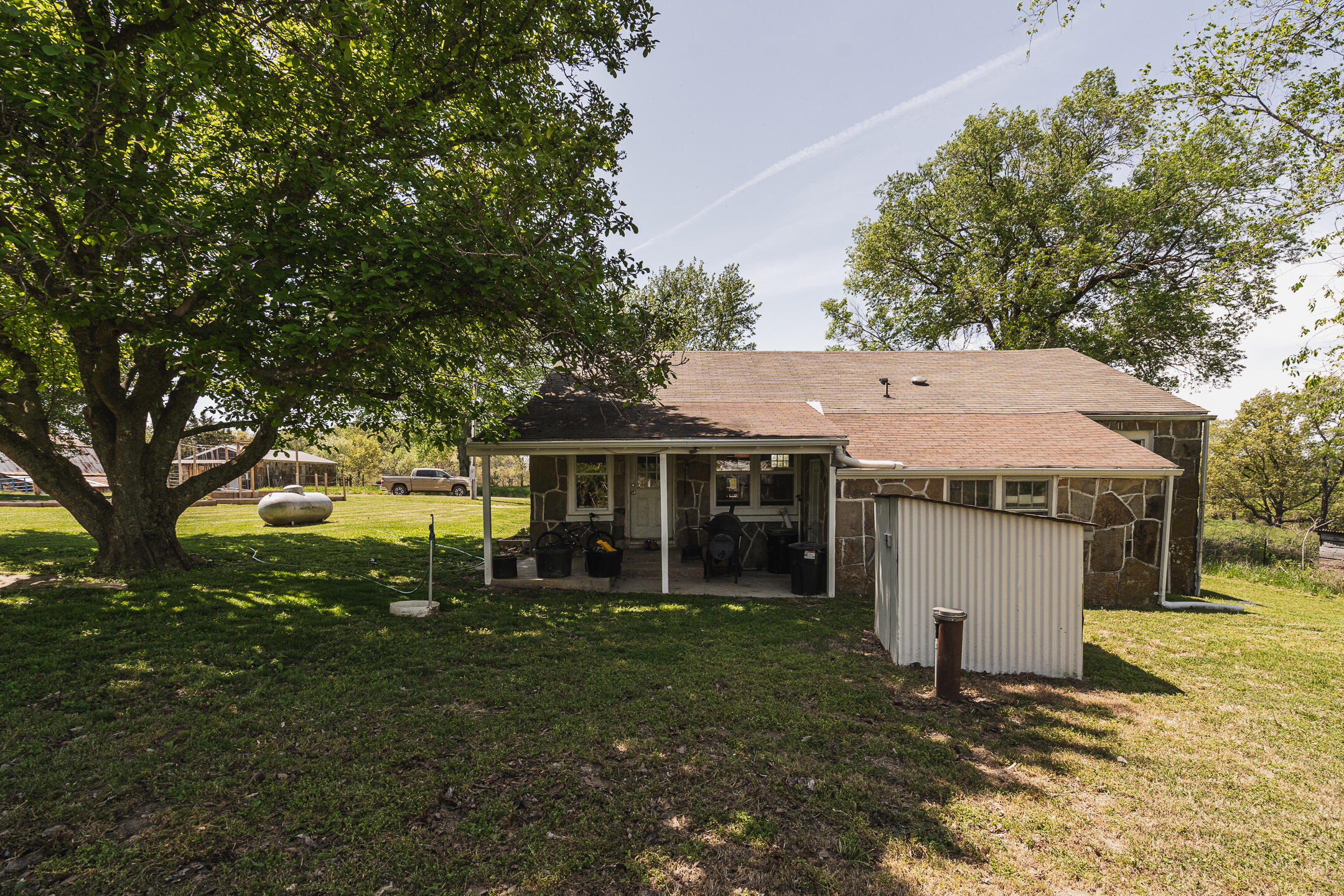 Stone house rear with a covered patio, small metal shed, and mature trees in the yard.