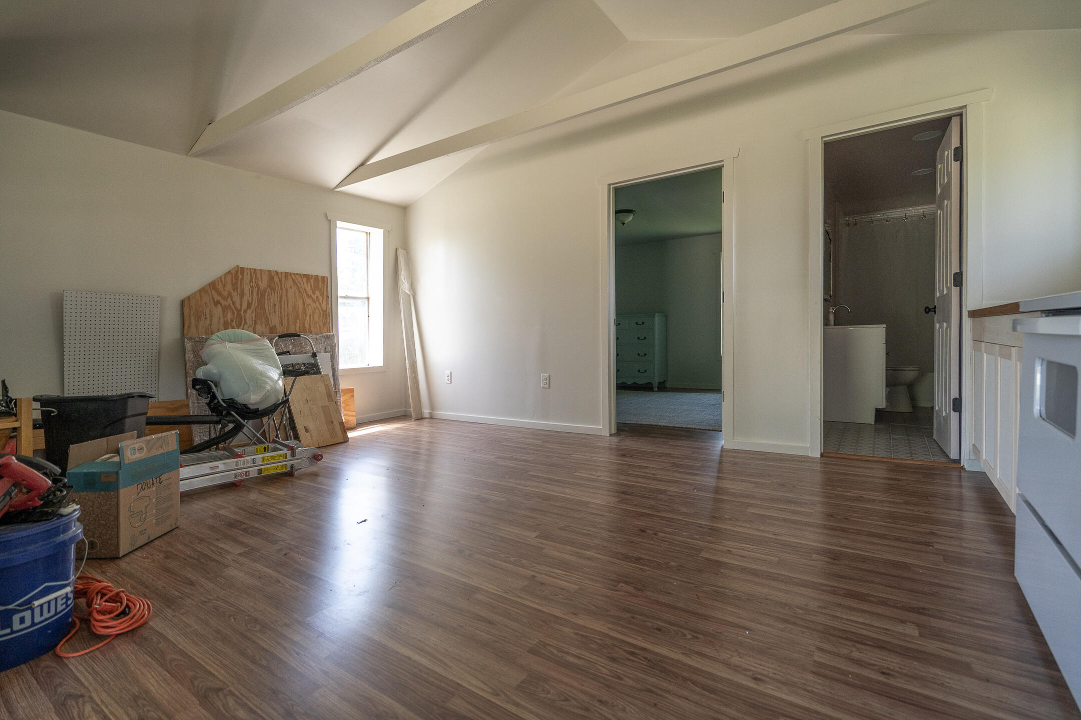 Vacant room with a vaulted ceiling, wood-look floors, kitchen cabinets, and materials piled beside the window.