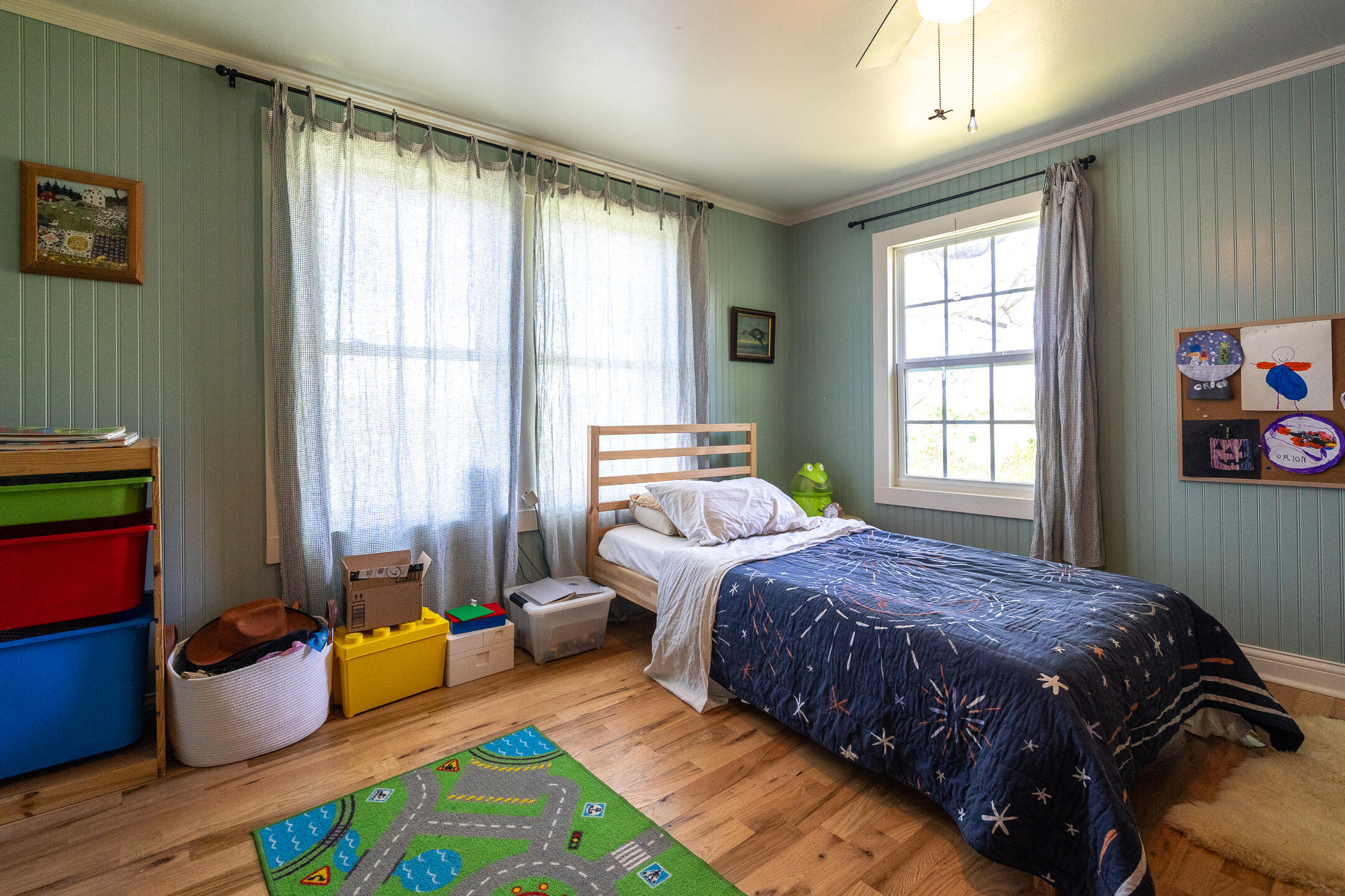 Bedroom with sage-green paneled walls, a star-patterned blue quilt, large windows, and a colorful road-play rug.