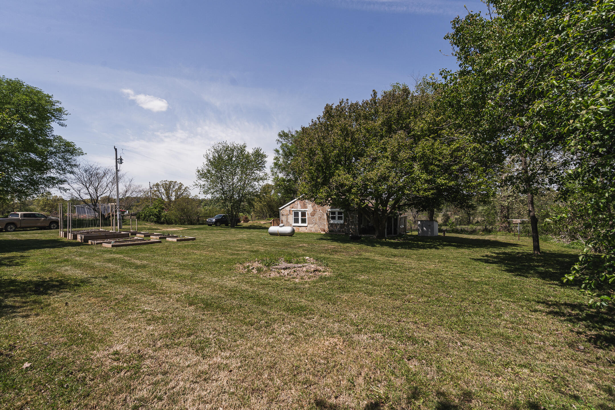 Small stone-clad house beneath mature trees, with raised garden beds and a propane tank in the yard.
