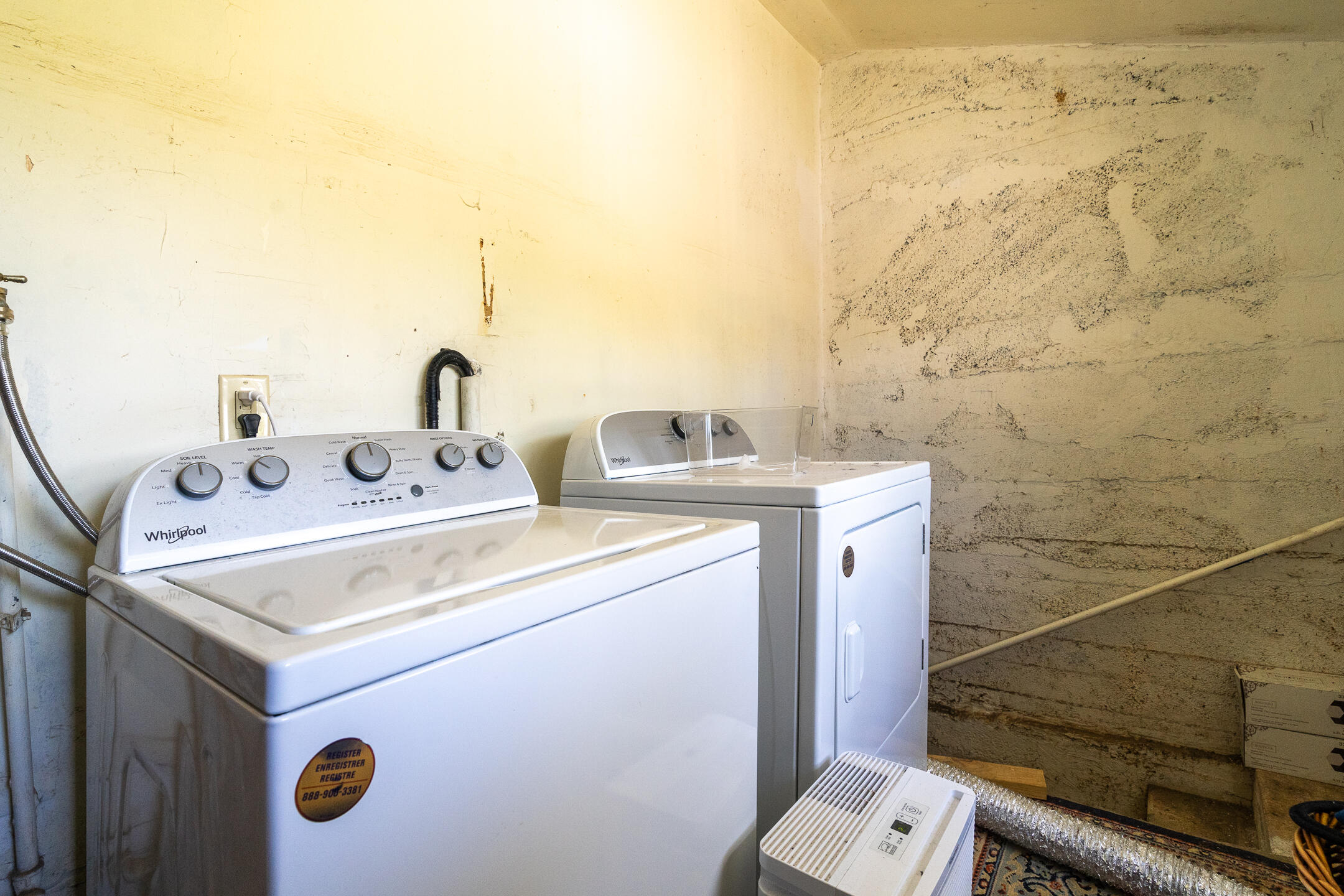 Laundry room with a top-loading washer, dryer, and portable air conditioner against unfinished walls.