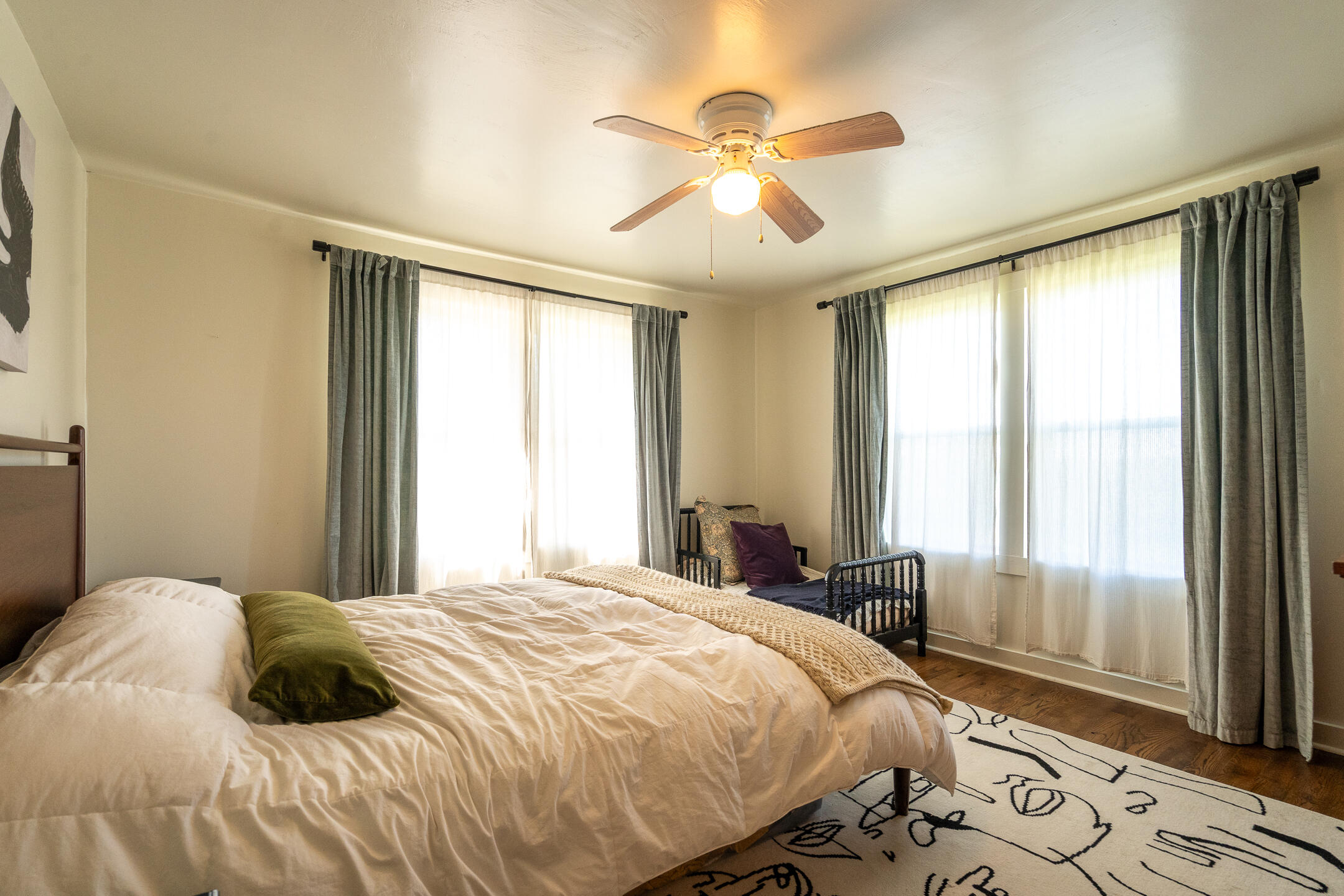 Bedroom with a large bed, small black crib, tall curtained windows, and ceiling fan.