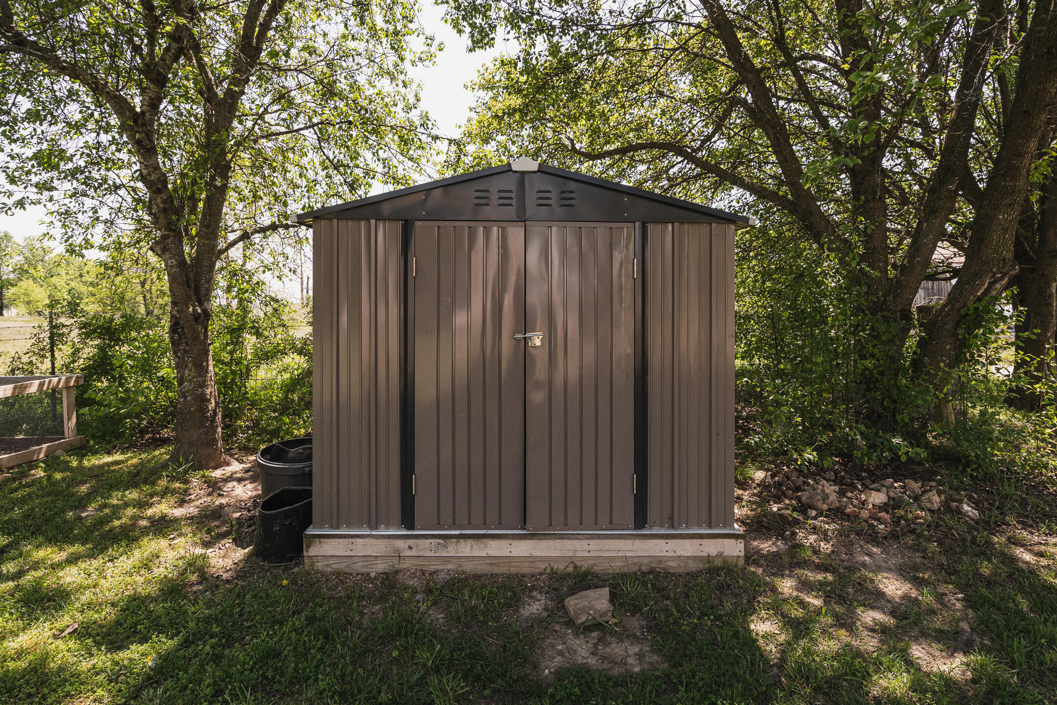 Brown metal storage shed with double doors beneath leafy trees.
