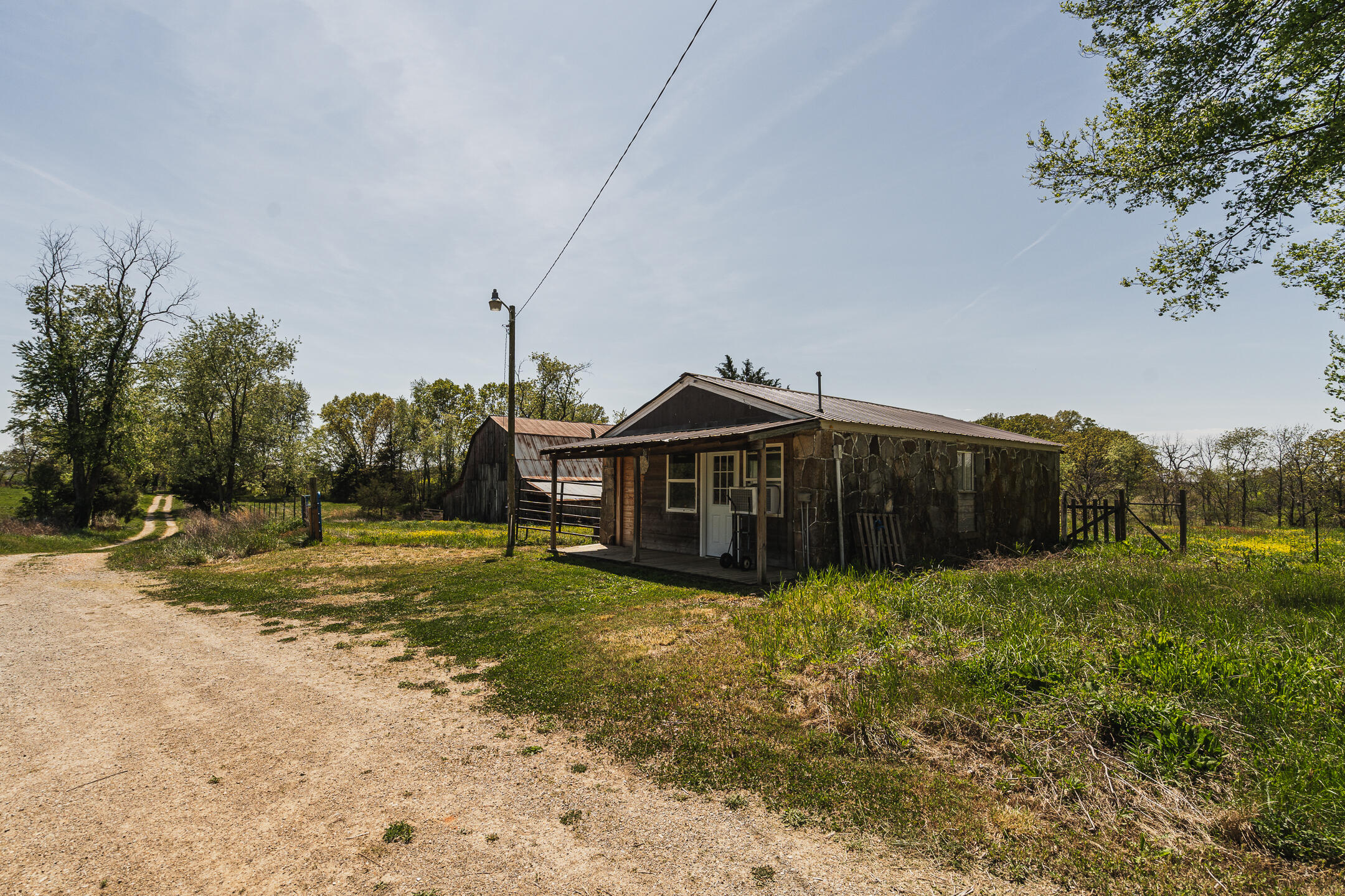 Rustic stone-and-wood building beside a gravel drive, with a metal-roofed barn behind it.