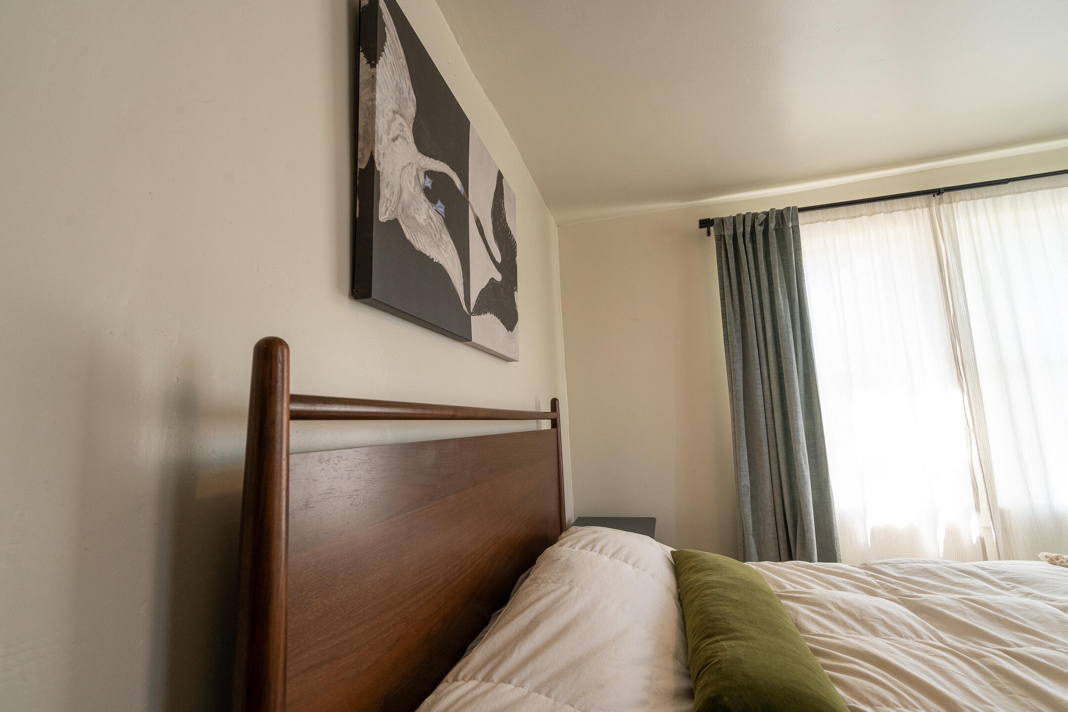 Bedroom with a dark wood headboard, green accent pillow, bird artwork, and gray curtains by a bright window.