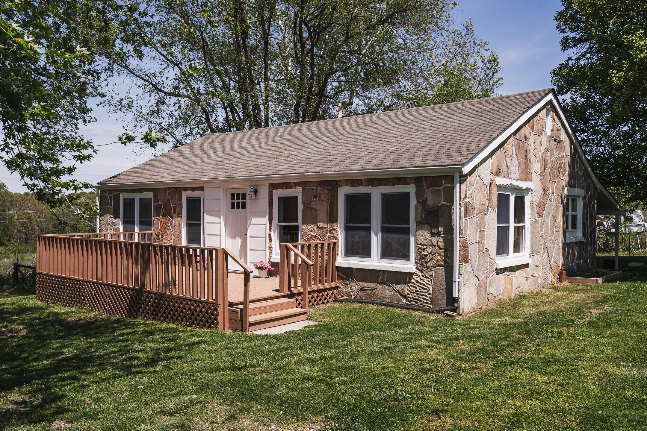 Single-story stone house with white trim and a broad wooden front deck beneath mature trees.
