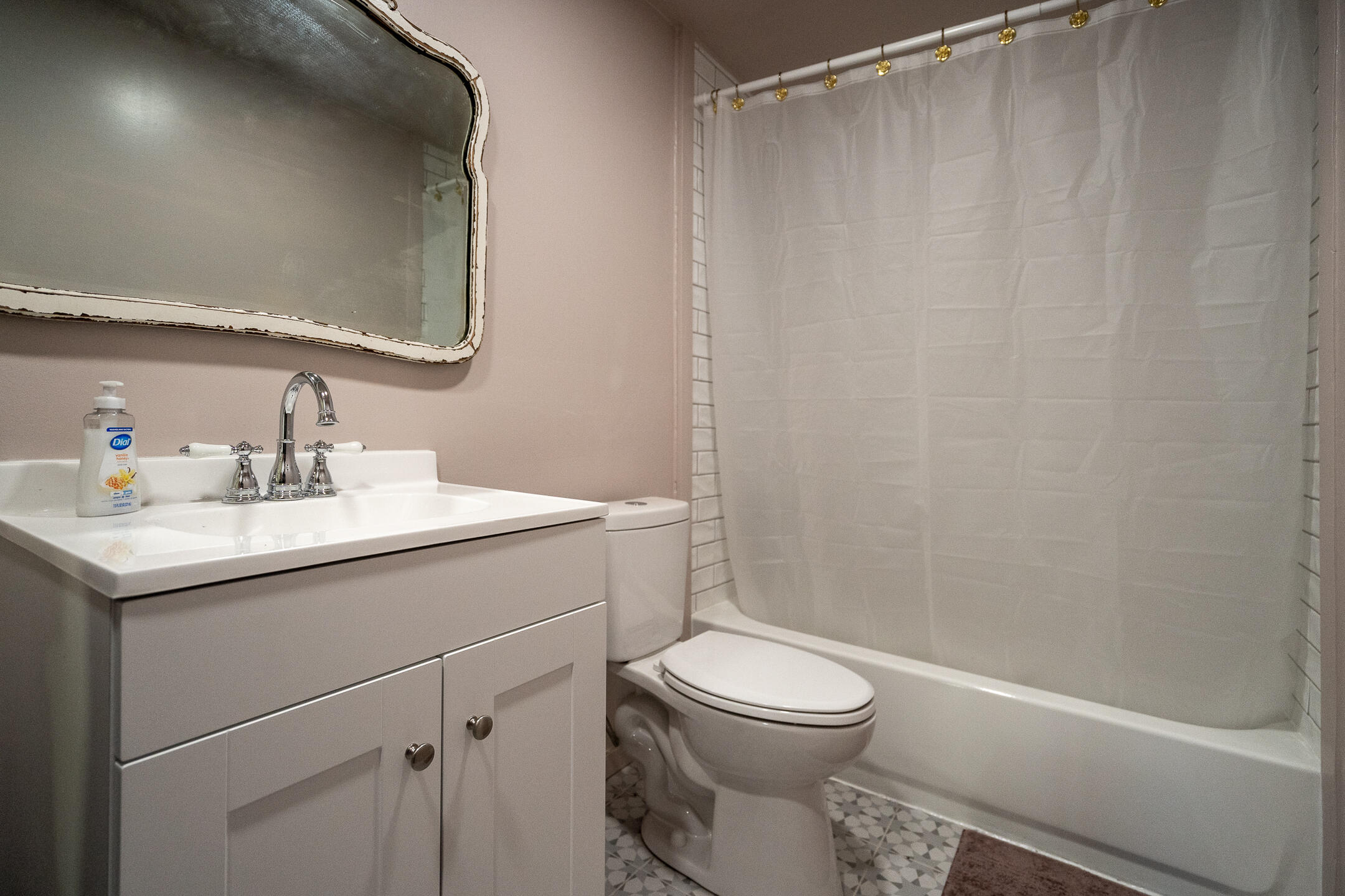Bathroom with white vanity, toilet, and bathtub, featuring a distressed-edge mirror and geometric tile floor.