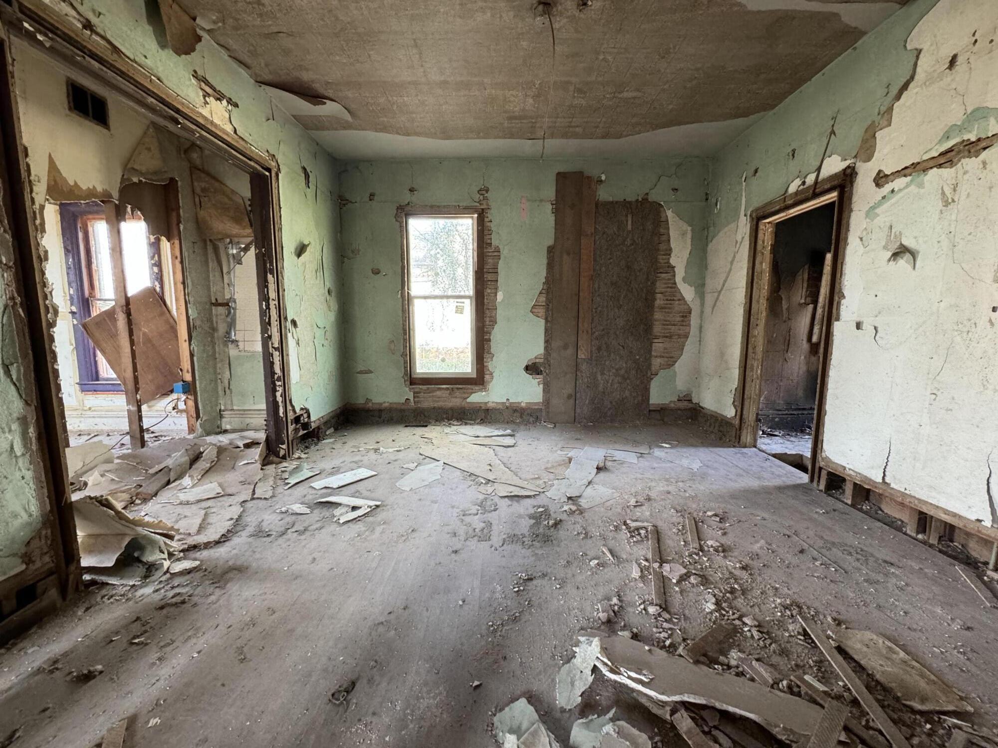 Vacant, damaged room with peeling green walls, exposed brick, a debris-covered floor, and a central window.