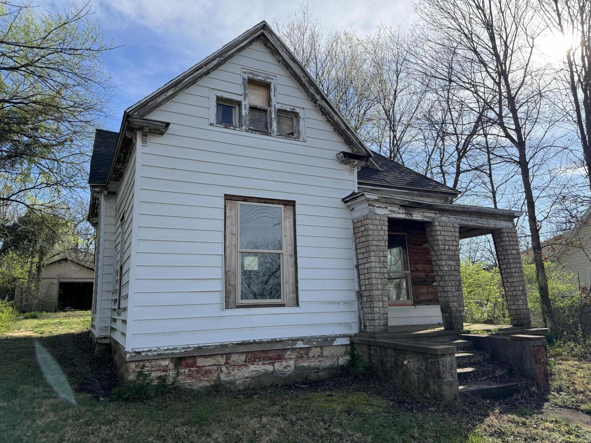 Weathered white two-story house with a steep gable and stone-column front porch.