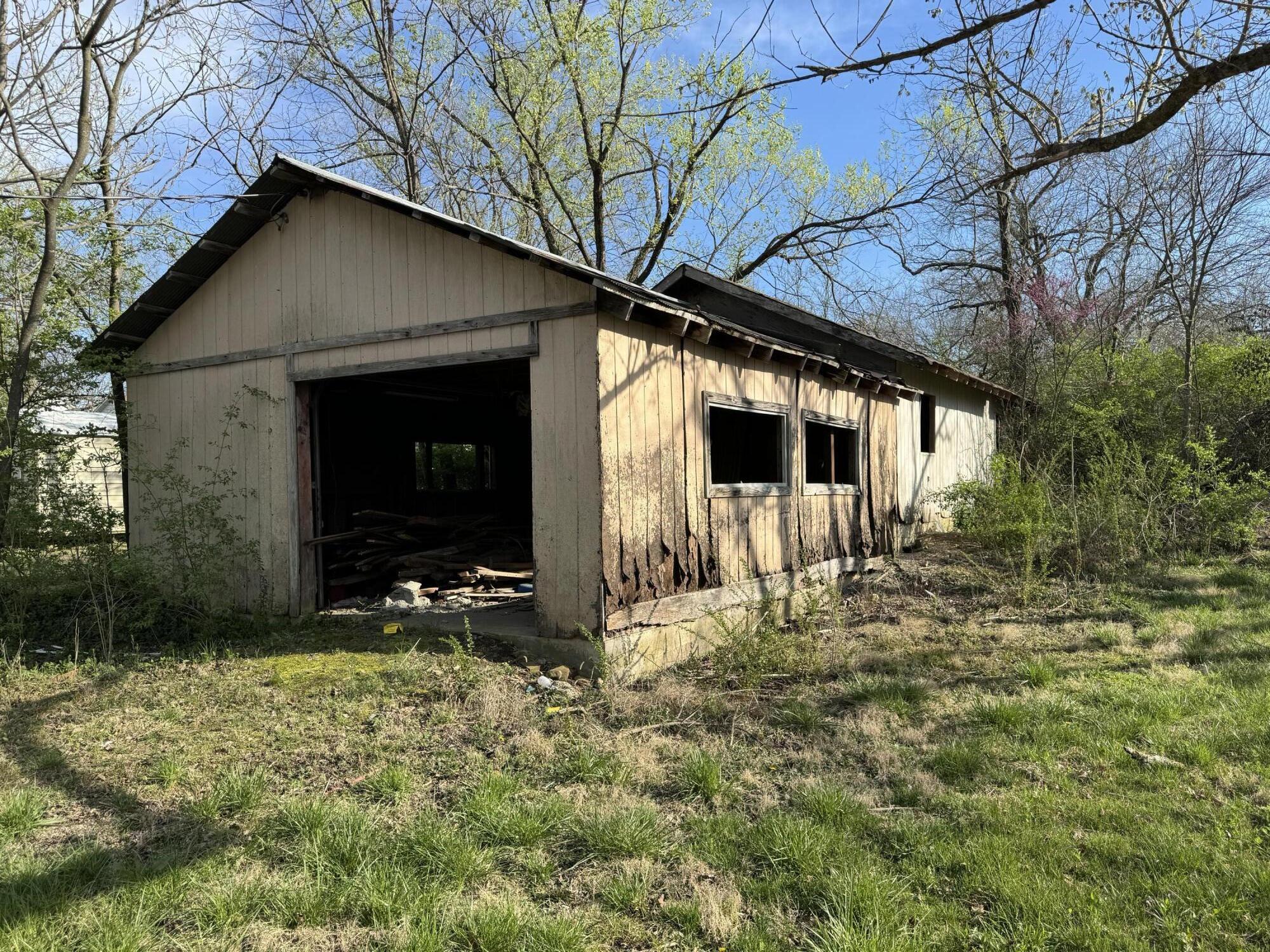 Weathered wooden outbuilding with an open doorway and damaged siding in a grassy, wooded yard.